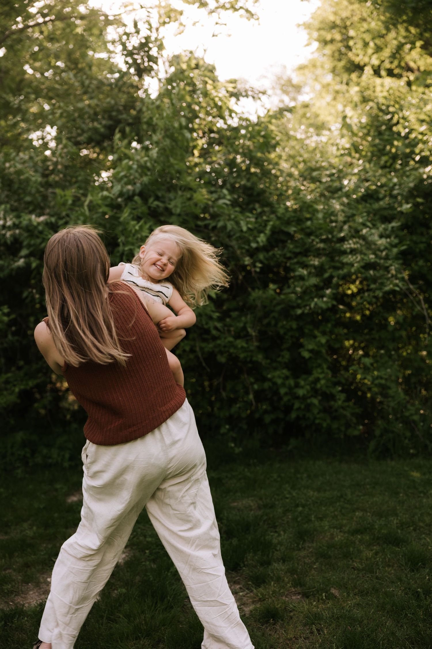 Mother joyfully spins her giggling toddler in a sunlit green garden.