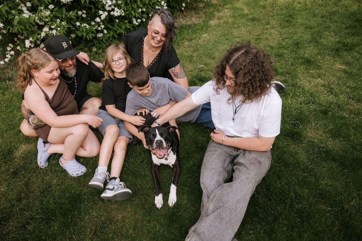 Family with children and a black and white dog sitting together on green grass outdoors near blooming bushes.