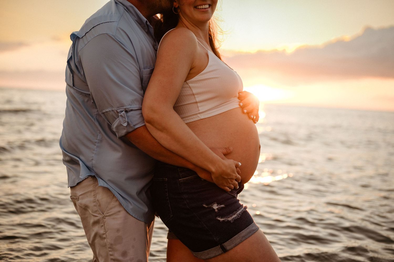 A pregnant couple embraces on the beach during golden hour sunset as waves roll in on the shoreline.