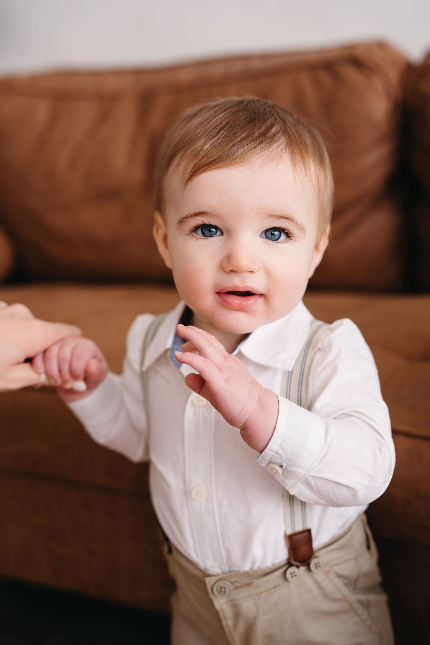 A small child in a white dress shirt and tan suspenders stands against a brown leather couch.