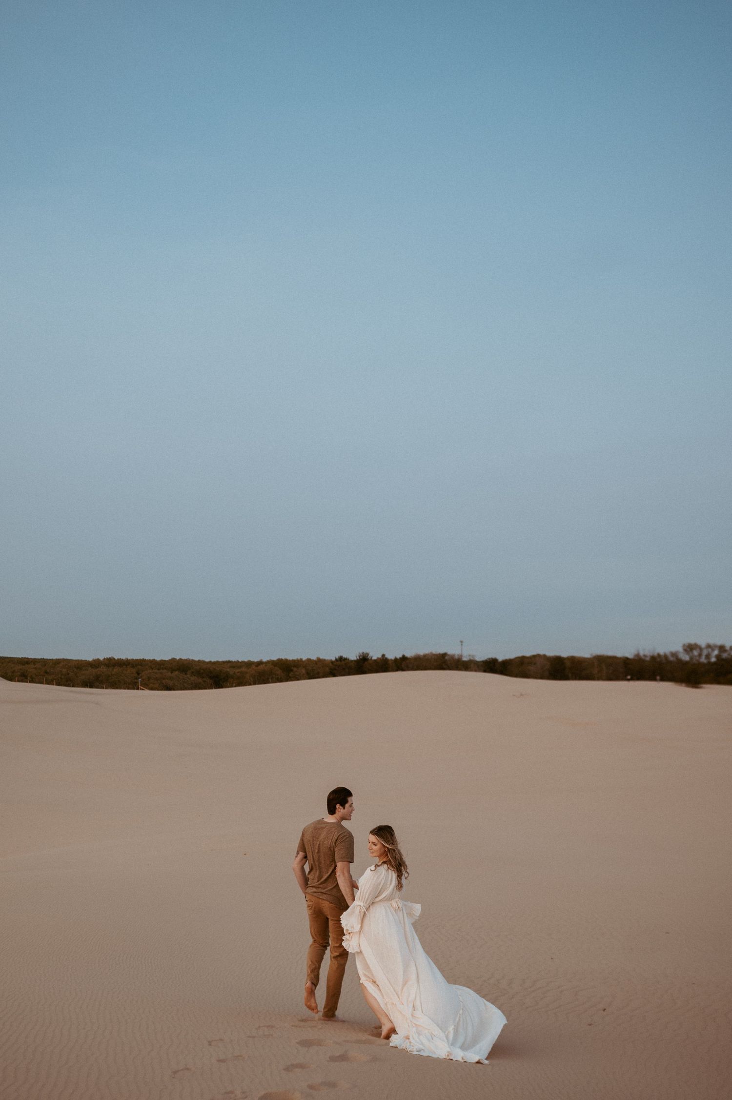 A couple walks up a desert sand dune during sunset photography session.