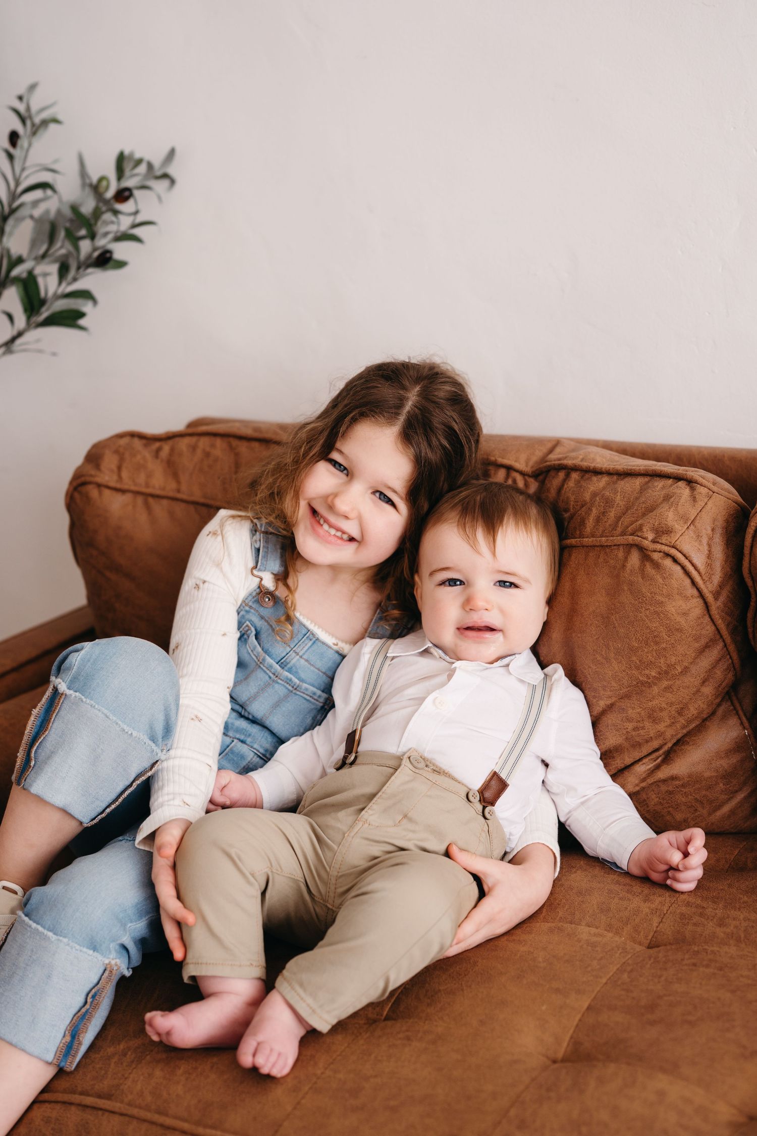 Two children sitting and smiling together on a brown leather couch with greenery in the background.