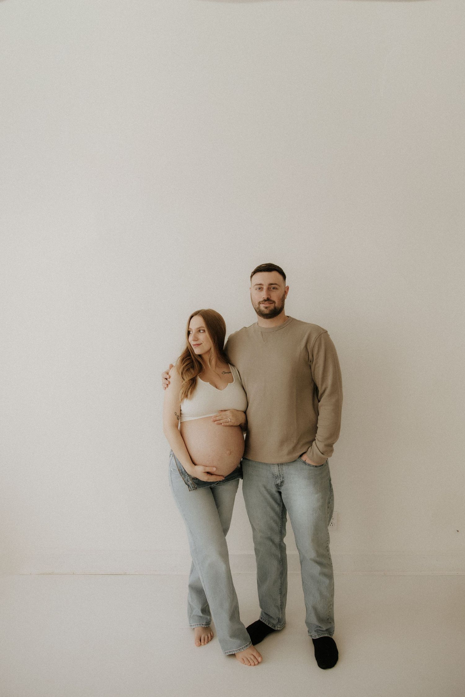 Expectant couple in matching neutral tones pose for simple studio portrait.