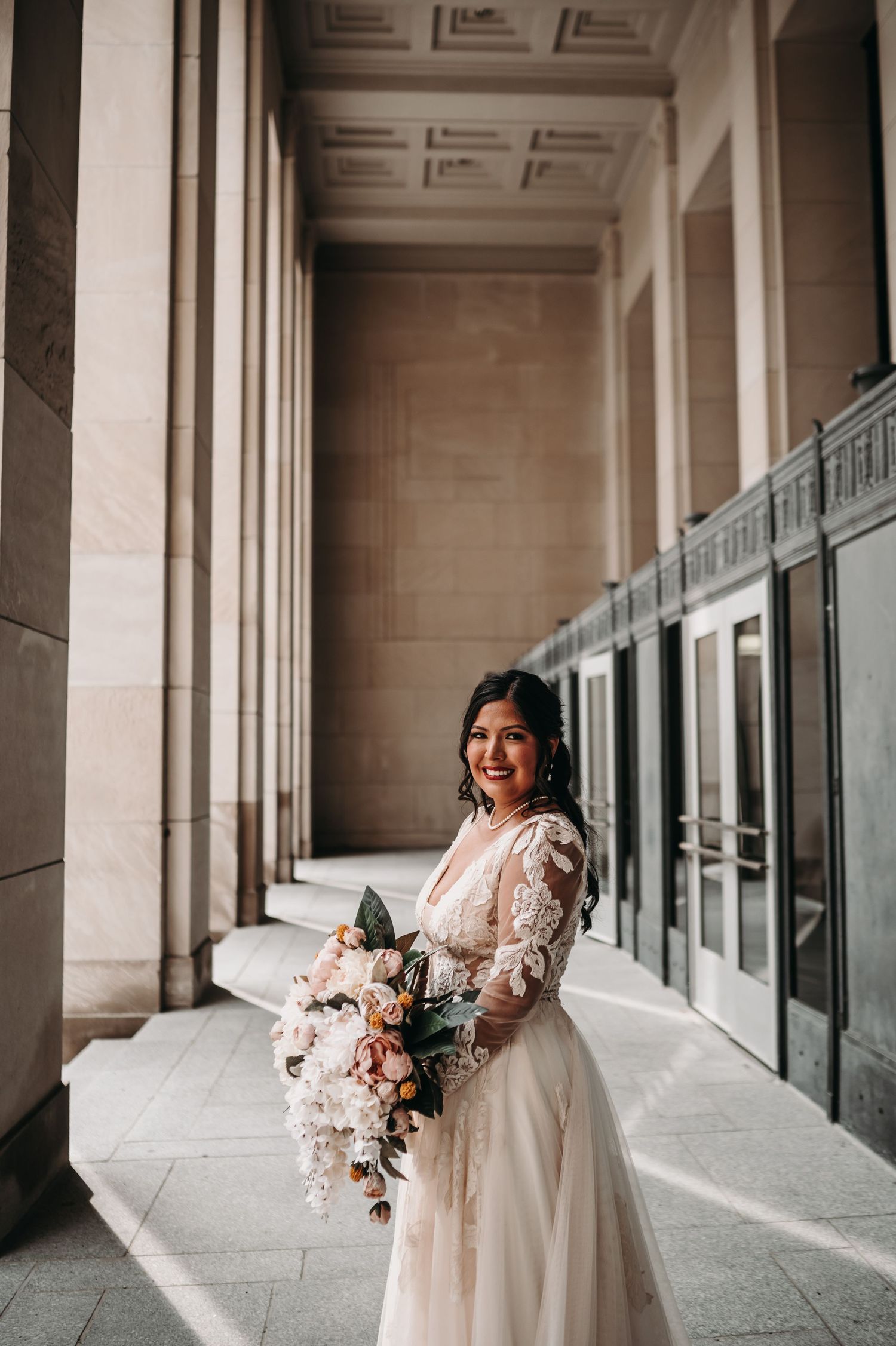 Bride in lace and tulle wedding gown holds blush bouquet while posing in elegant columned hallway.