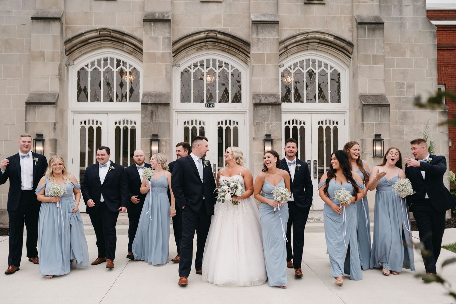 Wedding party in light blue bridesmaid dresses and black tuxedos poses in front of Gothic stone church entrance.