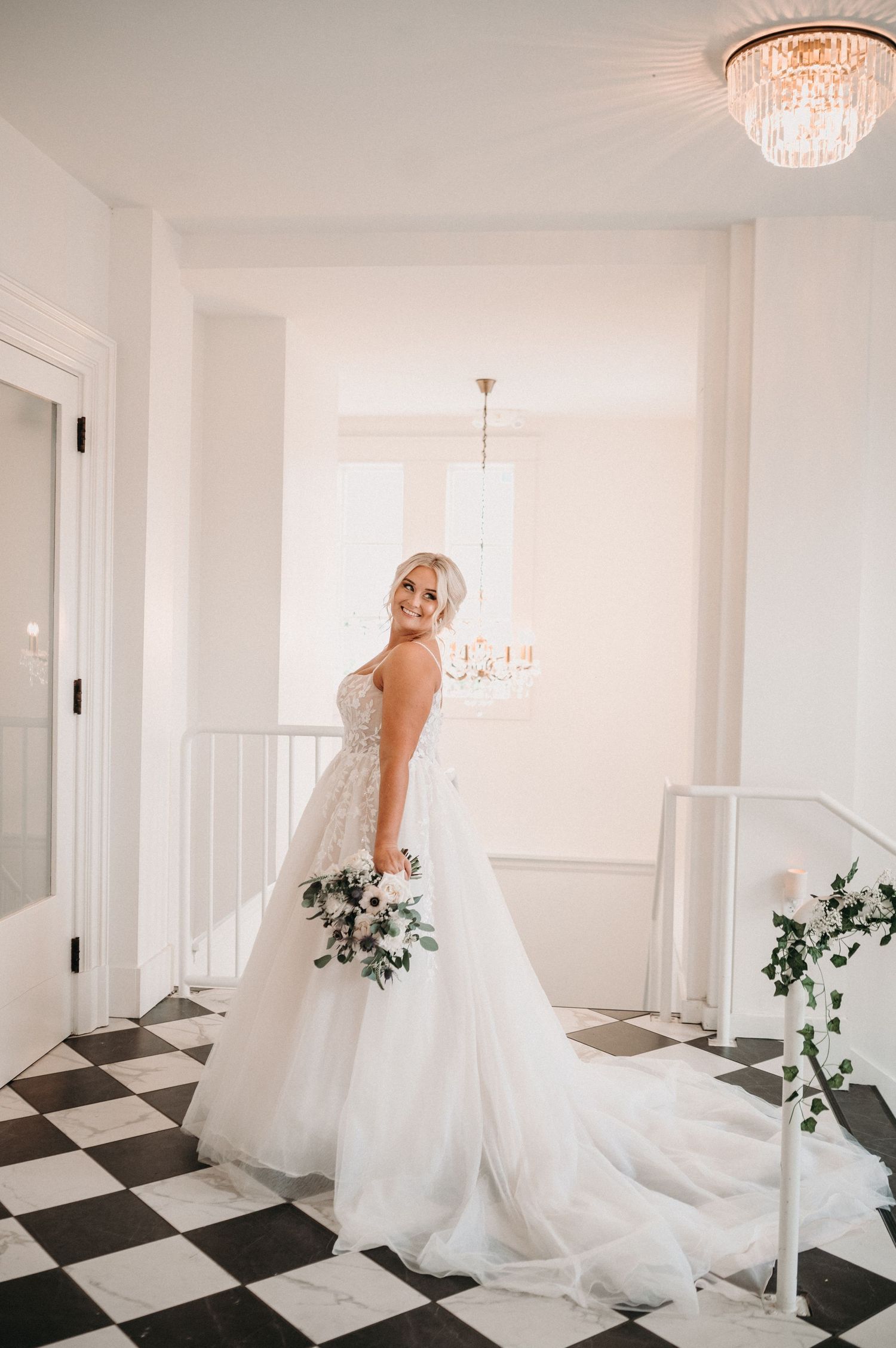 Elegant bride in flowing white gown poses in bright interior with chandelier and checkered floor.