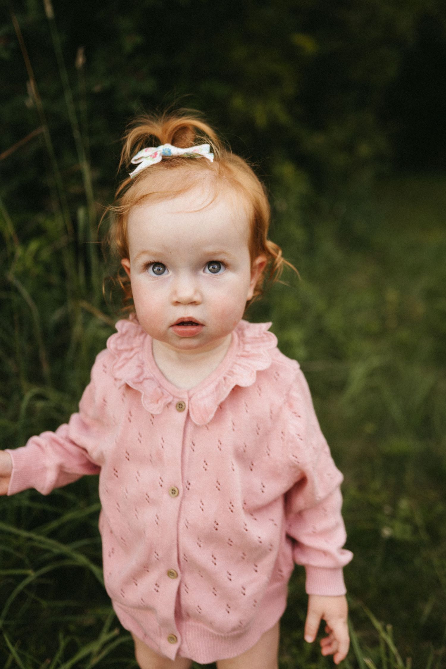 Small child wearing a pink eyelet cardigan outdoors against a dark green background.