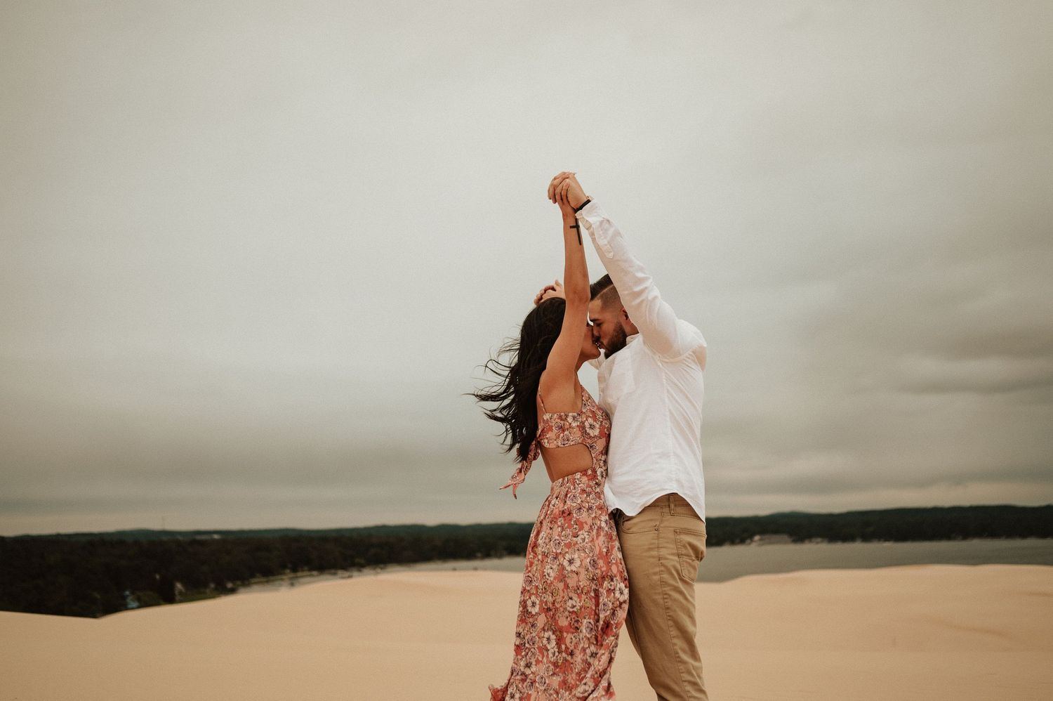 A romantic couple holds hands and leans back while dancing on sand dunes at sunset.