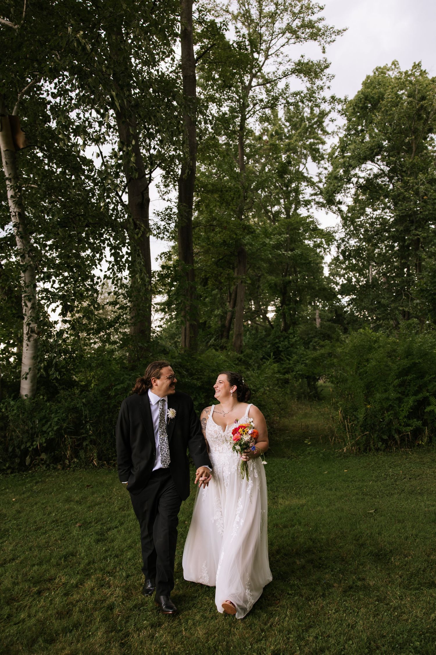 Couple in formal wedding attire walking together on a grassy hillside surrounded by trees.