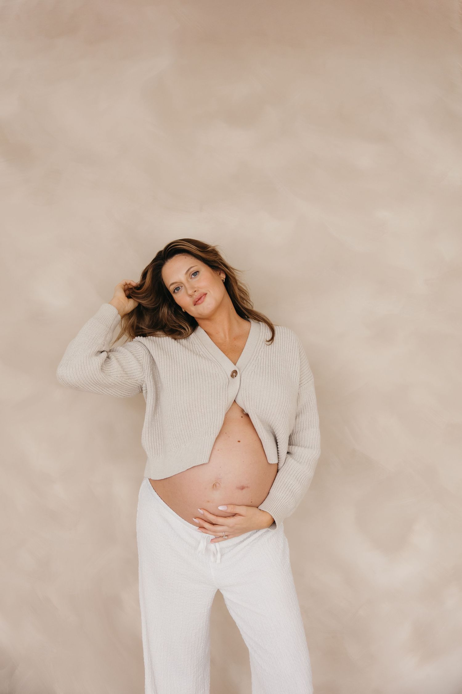 Smiling pregnant woman in beige cardigan and white pants cradling her bump against a neutral wall.