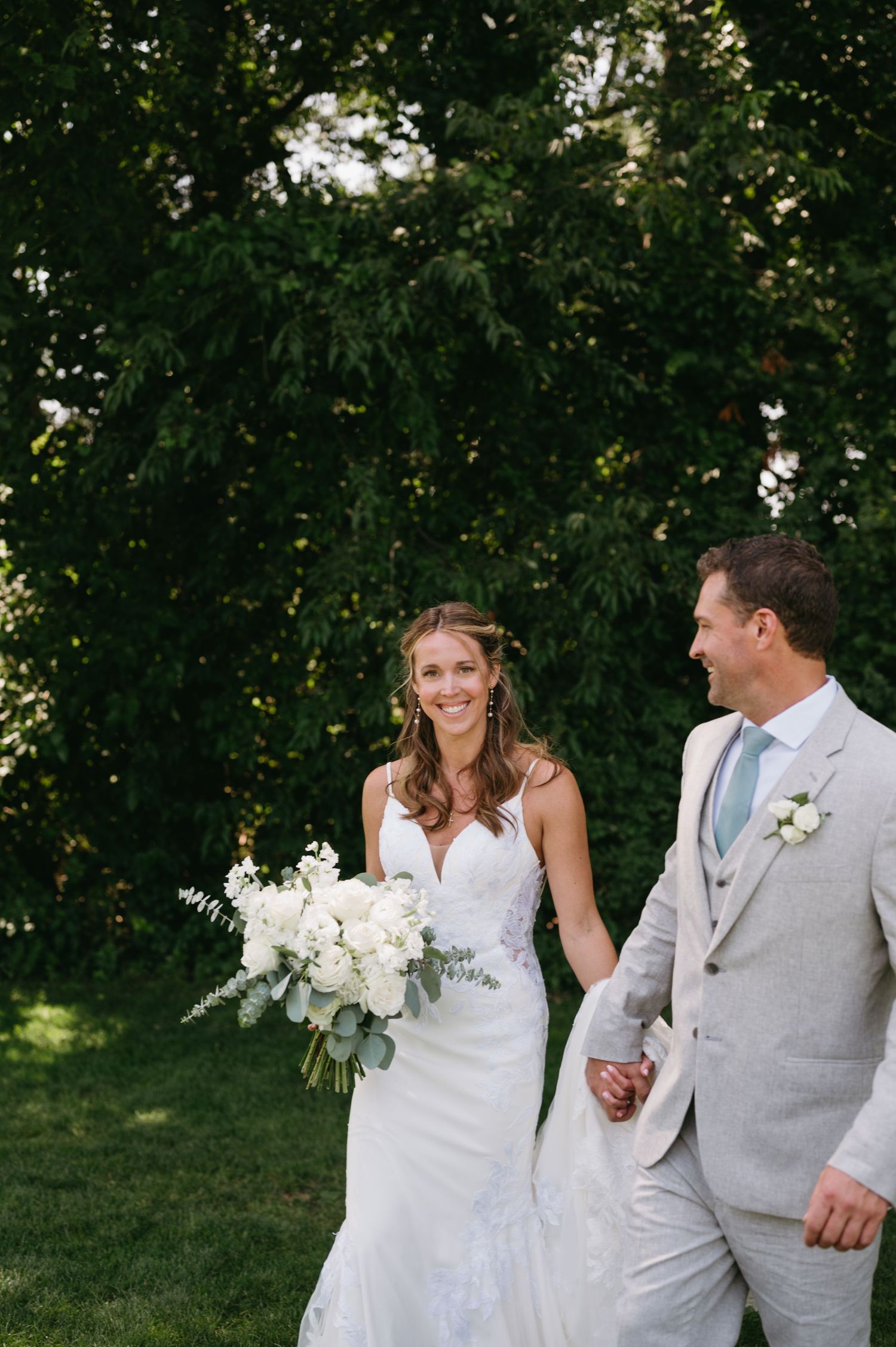 A couple in wedding attire walking together on a sunny lawn with trees in the background.