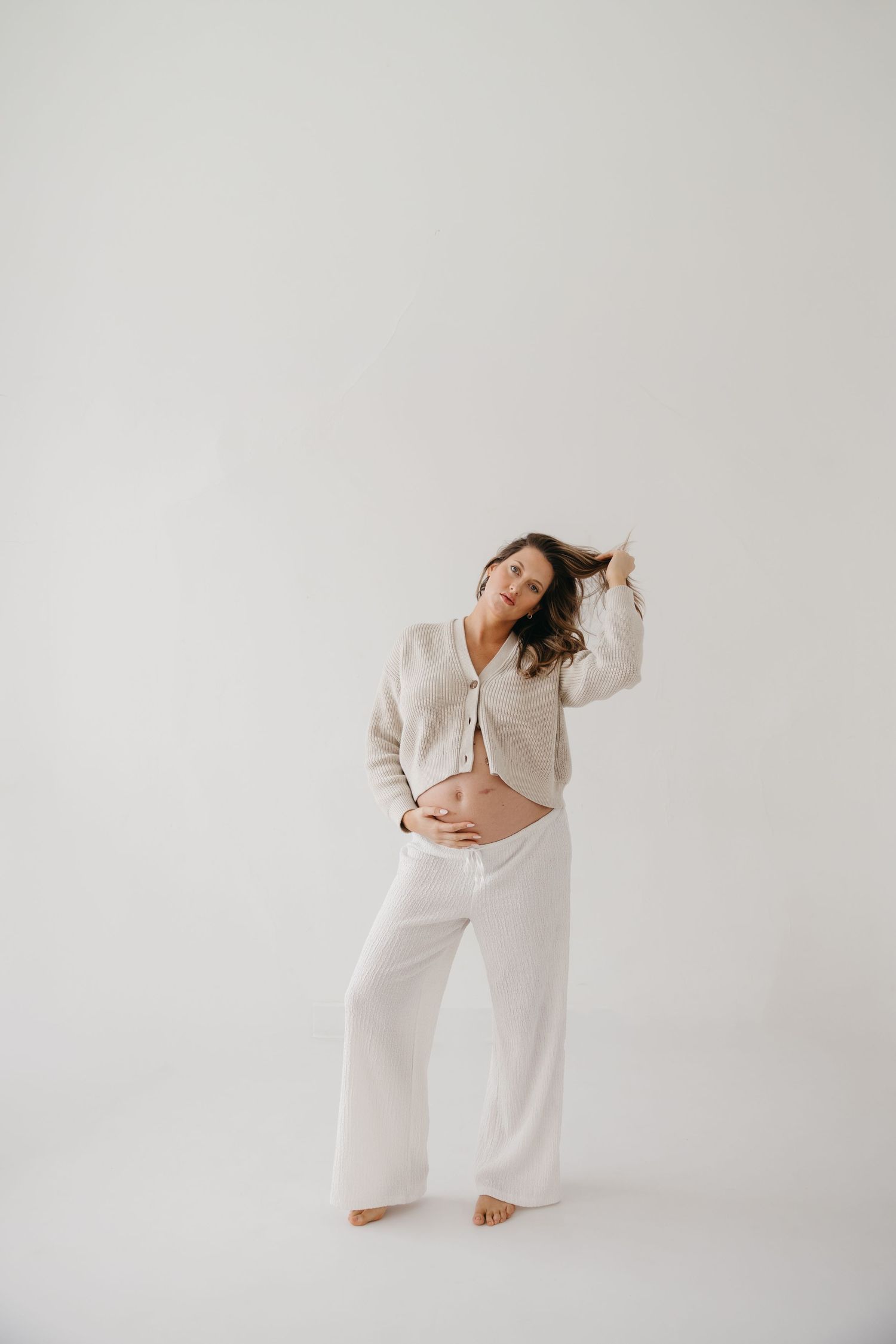 Pregnant woman in beige cardigan and white pants cradling baby bump, posing barefoot against white background.