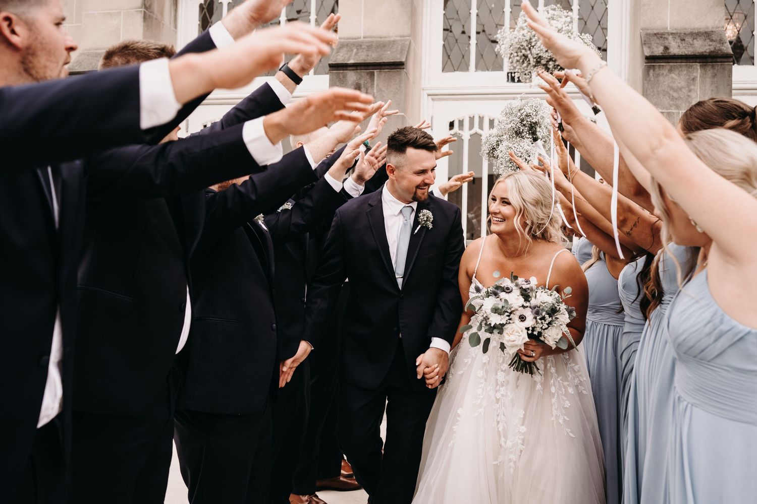 Wedding party celebrates with raised hands in a joyful moment outside a church entrance.