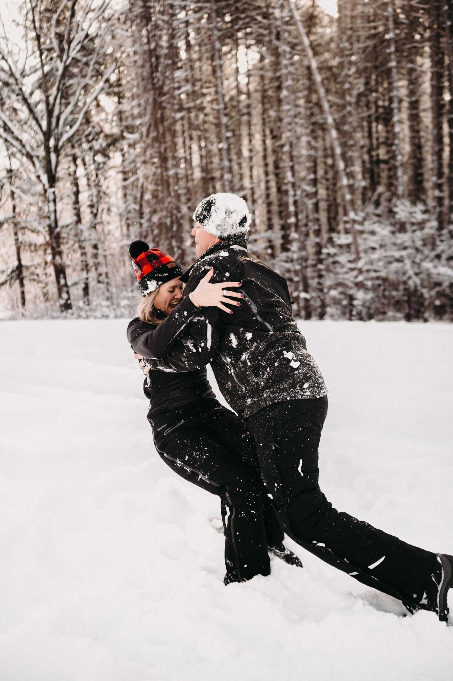 A couple embracing in winter attire against a snowy forest backdrop.