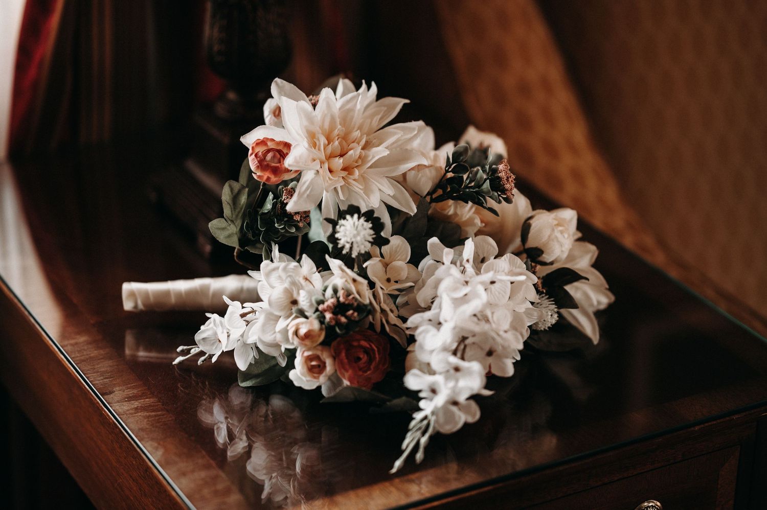A delicate bridal bouquet with white and blush flowers rests on a dark wooden surface in moody lighting.