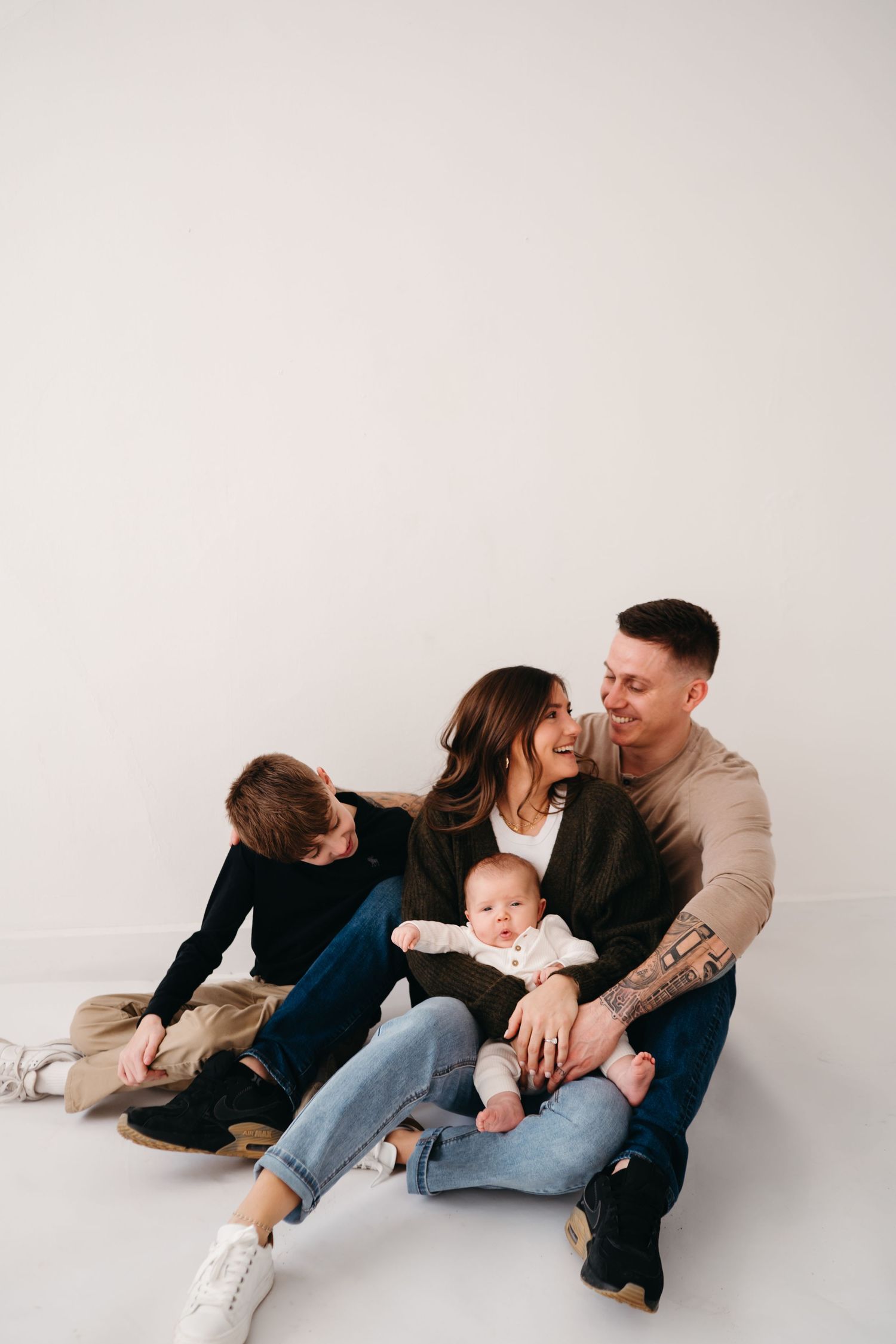 Intimate family portrait with parents relaxing on floor with baby and older child in casual clothing.
