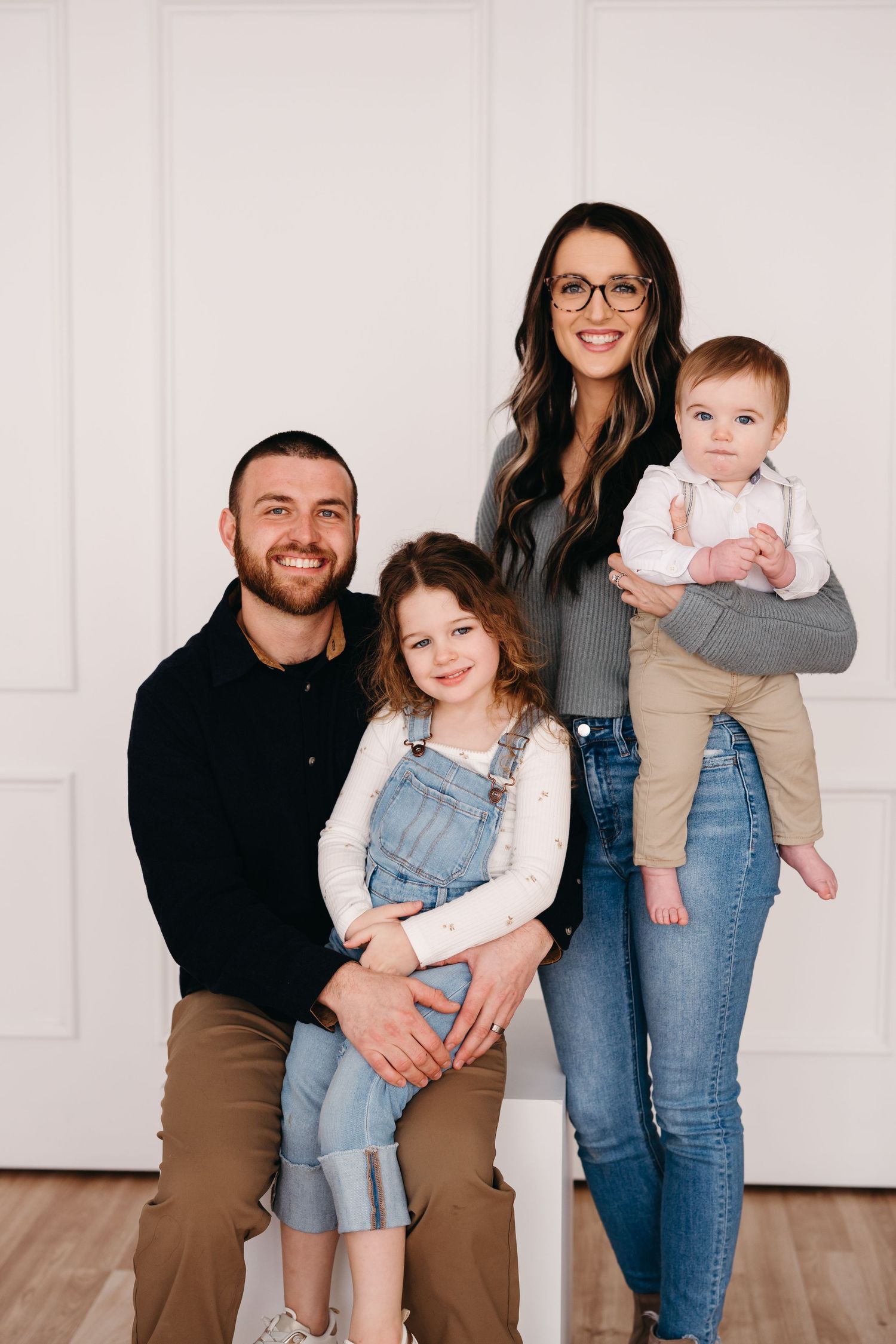 A family of four poses together against a white wall wearing coordinated casual outfits.