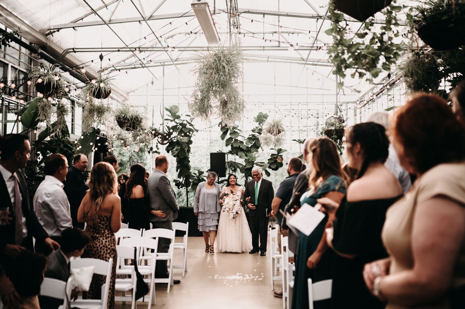 Wedding ceremony in a glass greenhouse filled with plants and guests seated in white chairs.