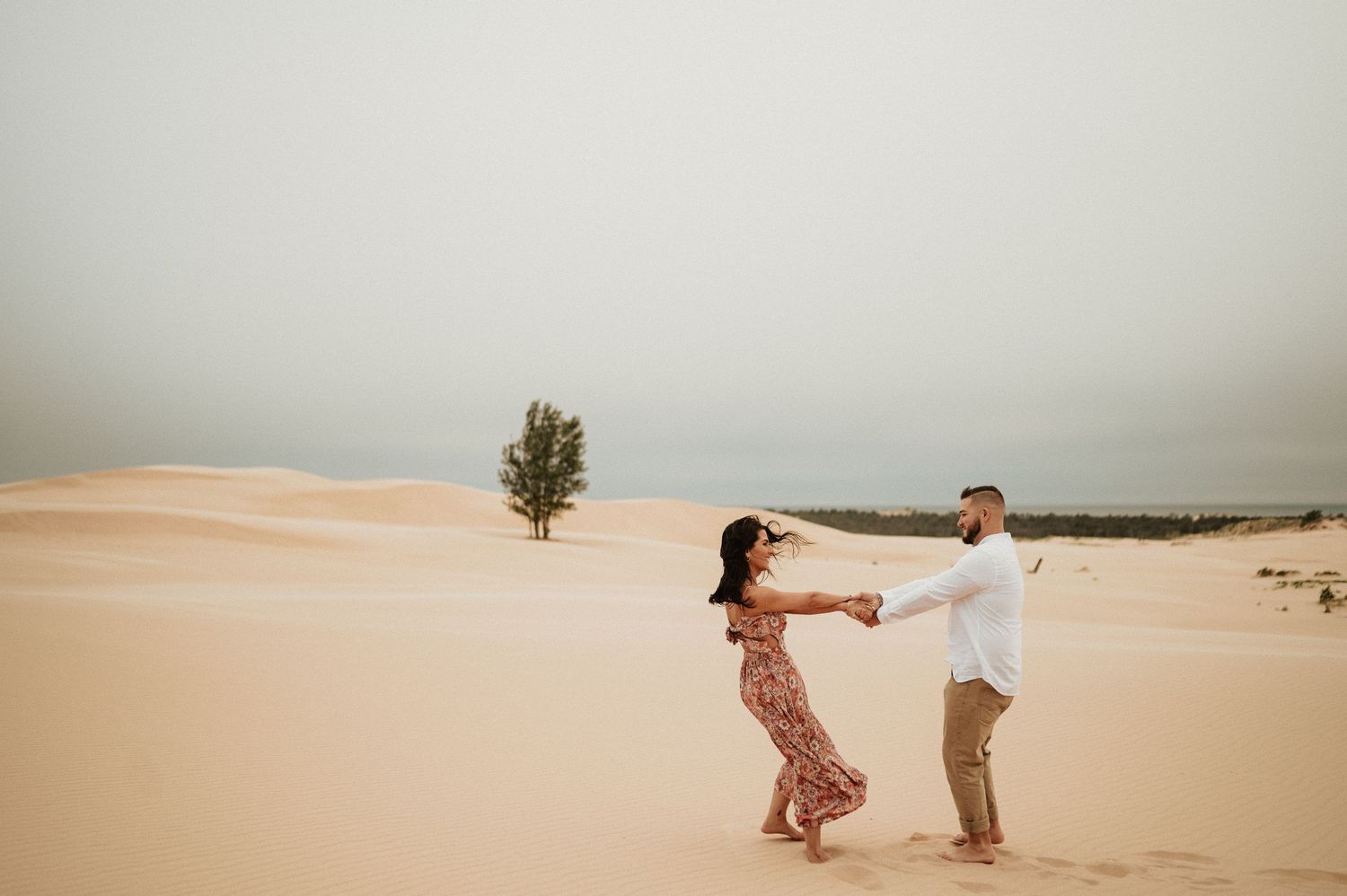 A couple dances together on sand dunes with a lone tree in the background under overcast skies.
