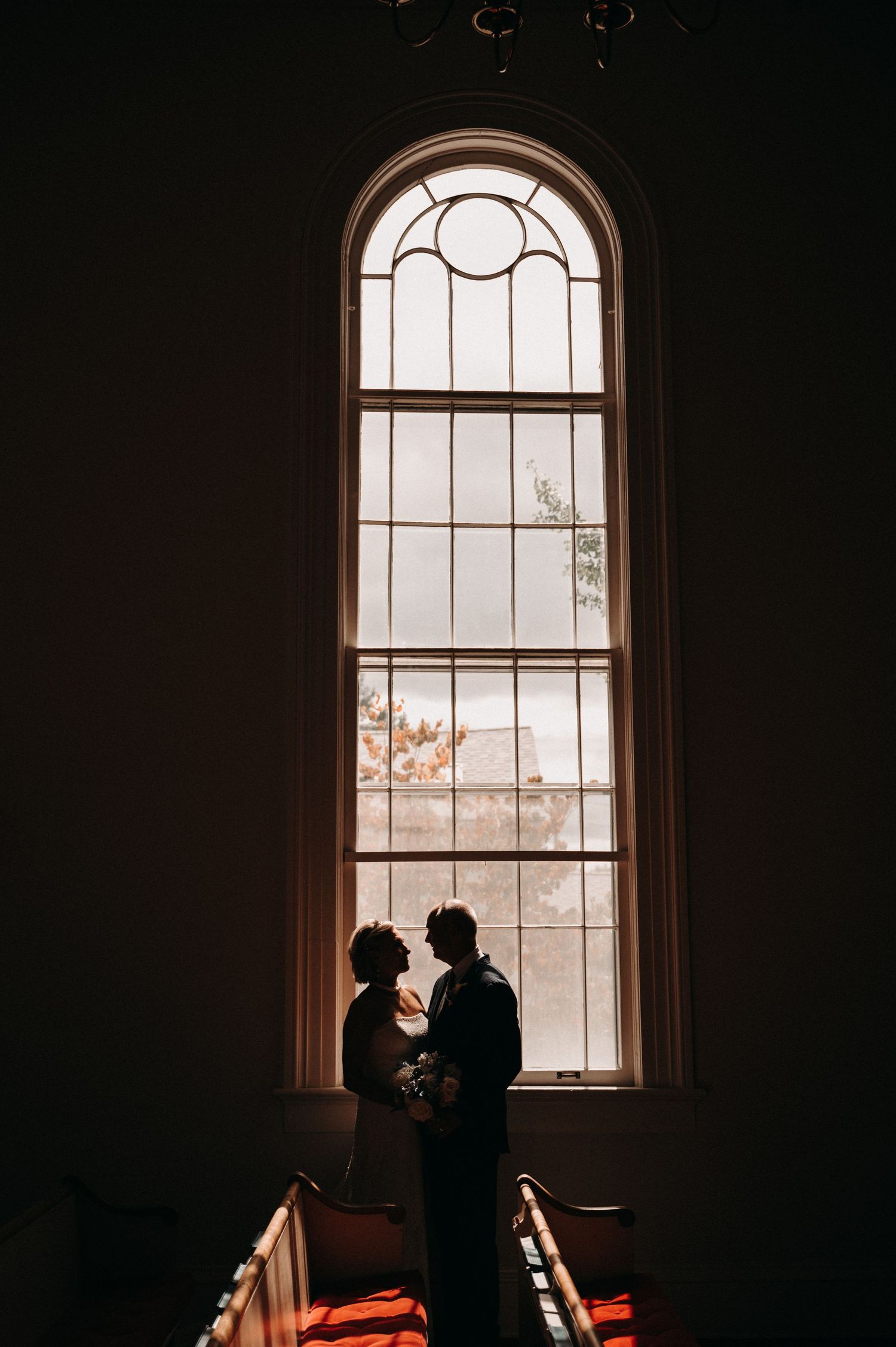 Silhouetted couple stands in romantic pose by arched window in dark interior.