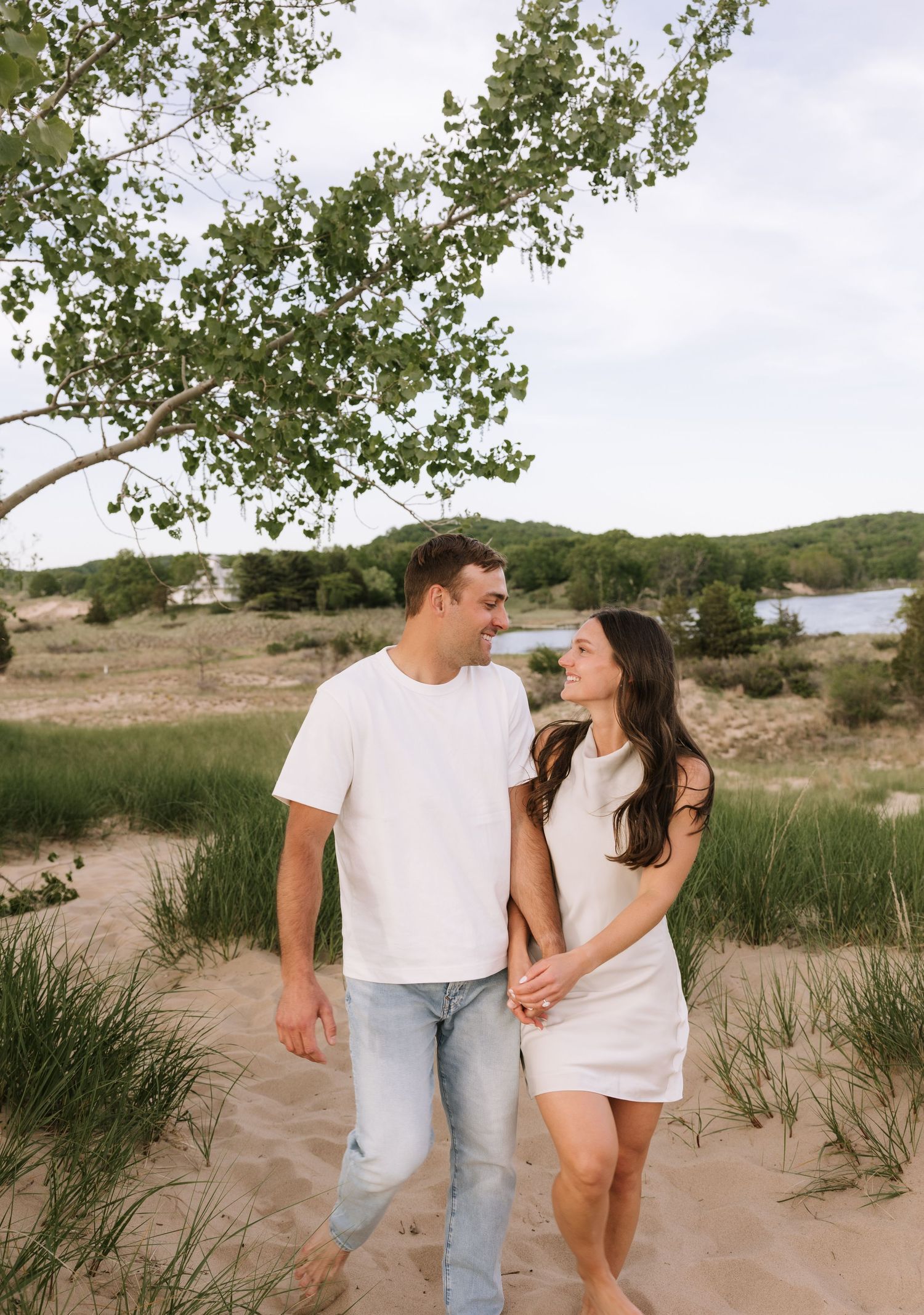 Couple holding hands and smiling at each other while walking barefoot on sandy dunes near water.