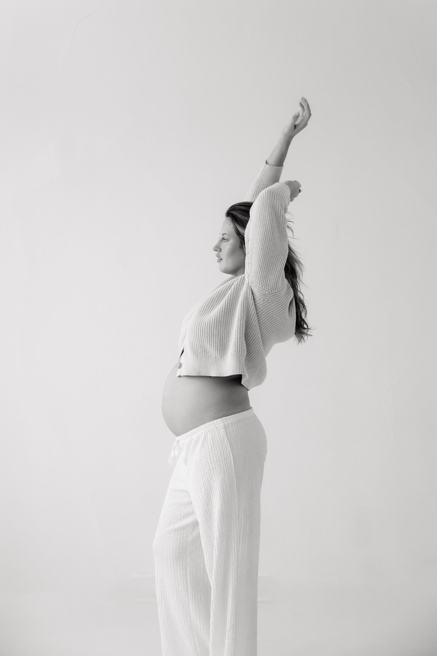 Black and white photo of pregnant woman stretching arm overhead in white studio setting.