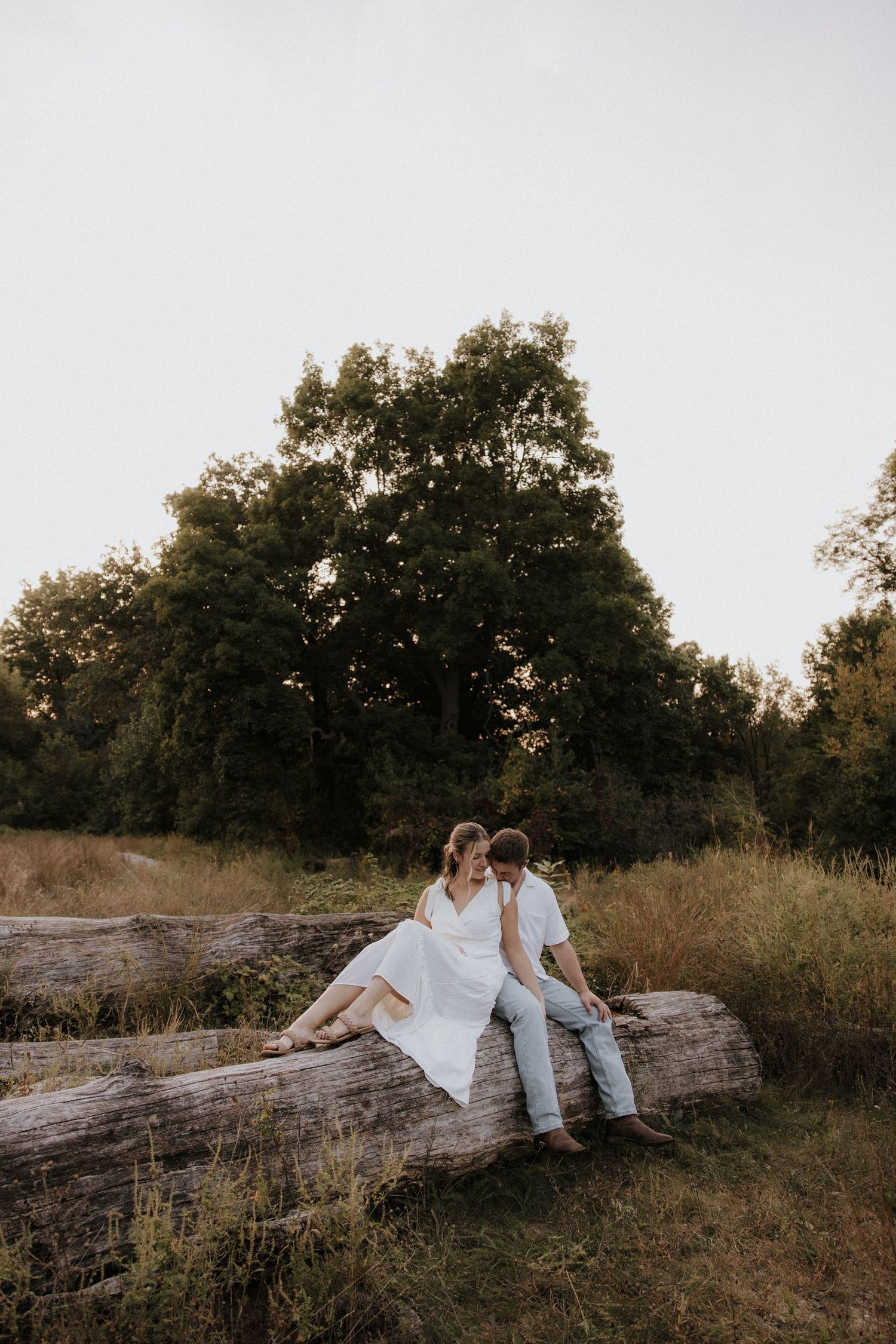 Couple dressed in white sits together on fallen log in natural outdoor setting at sunset.