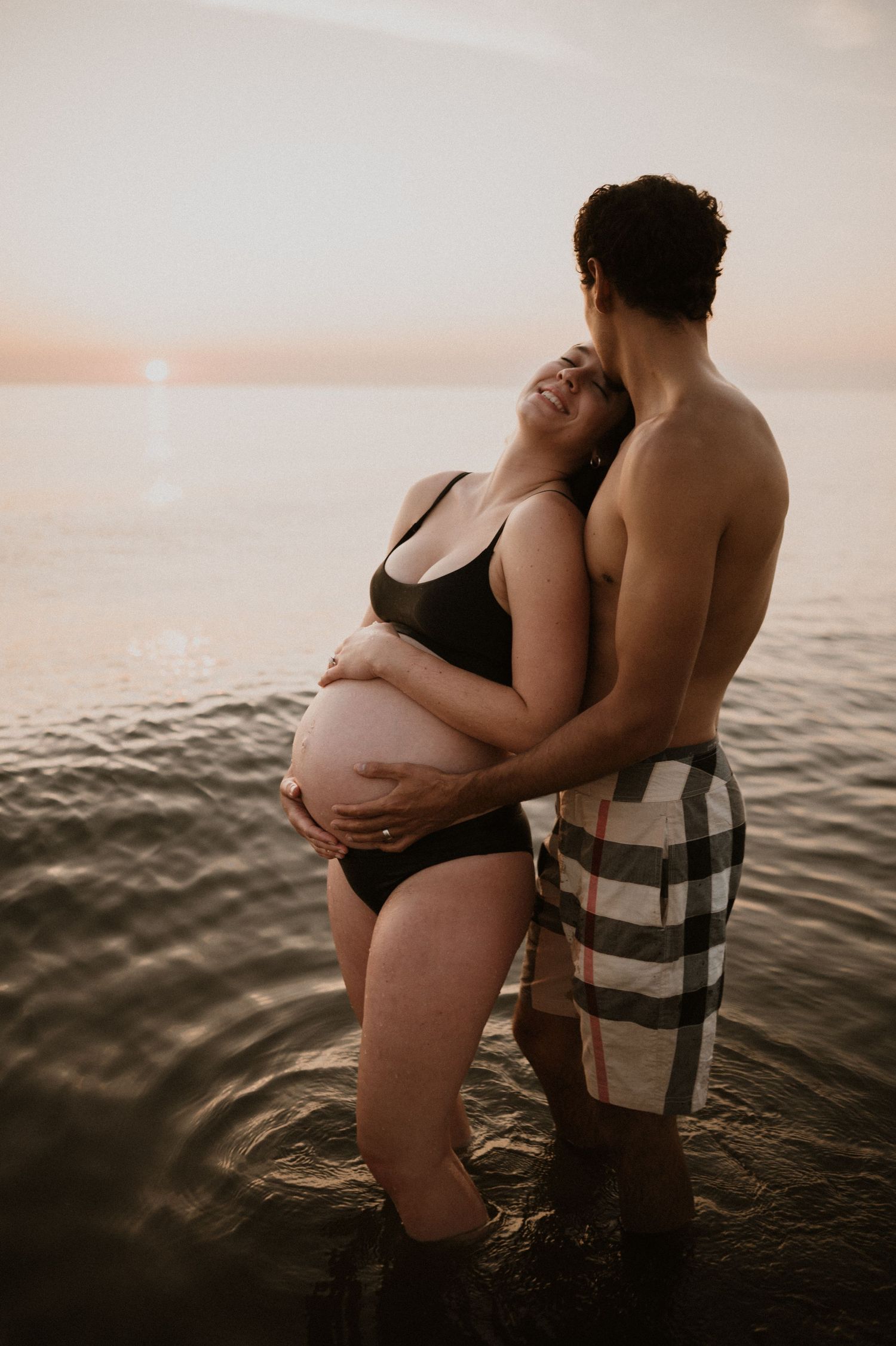 Intimate maternity moment captured with couple embracing in ocean water at sunset.