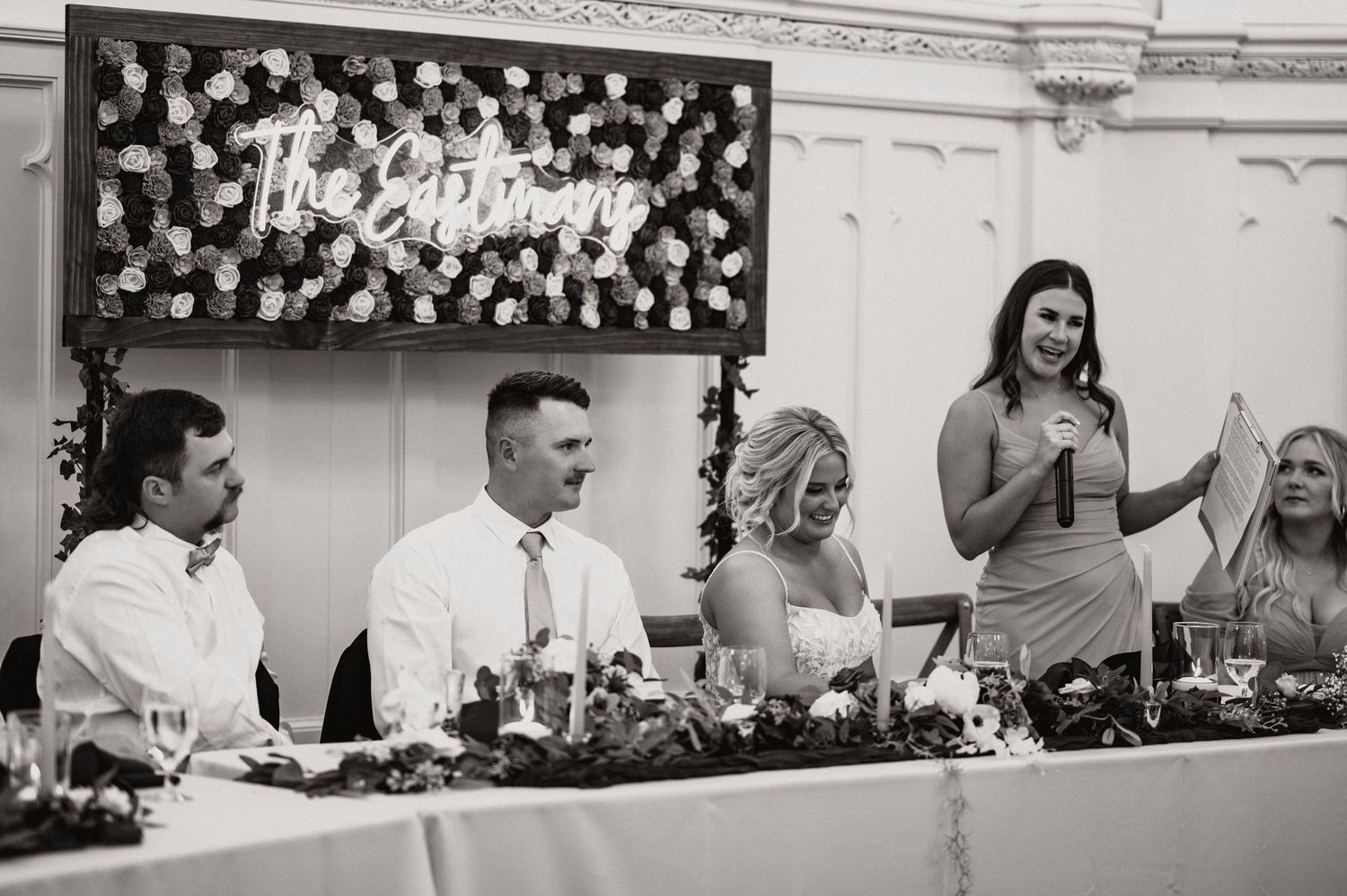Black and white photo of wedding reception speeches at head table with floral backdrop.