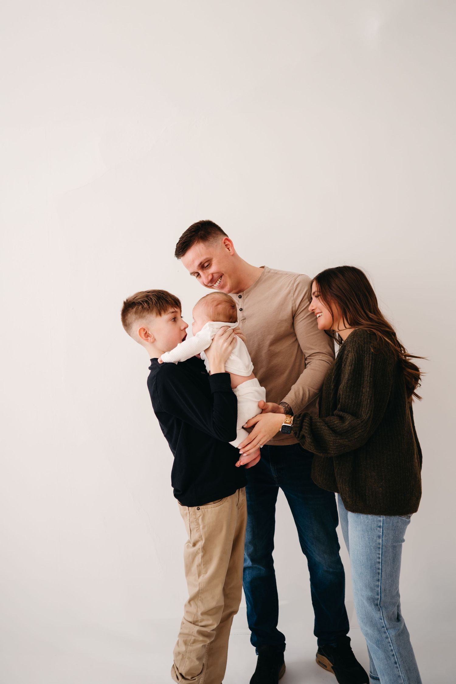 Family of four standing together as they admire their newborn baby against a light backdrop.