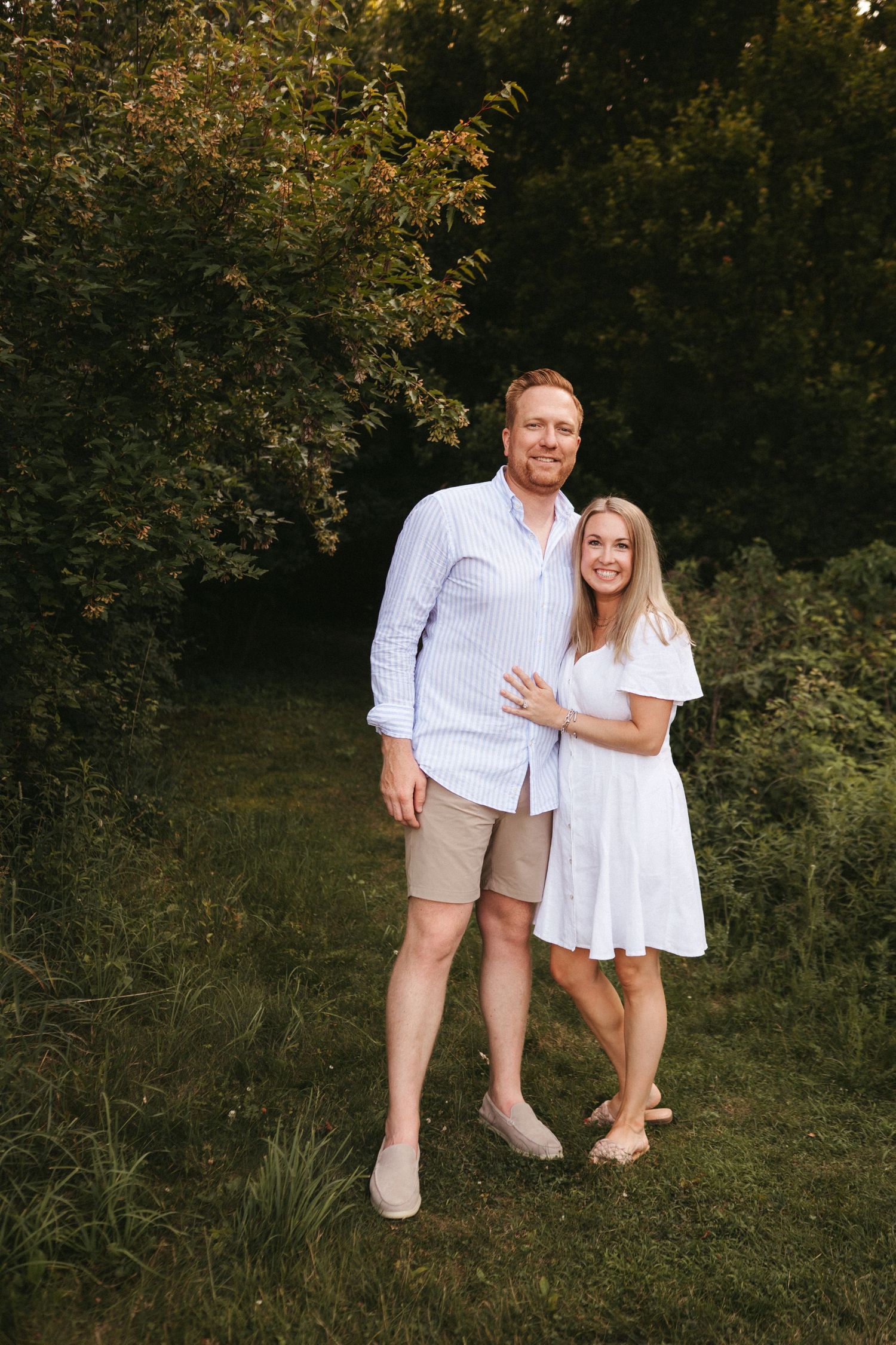A couple in coordinating white and khaki outfits stands together on grass with trees in background.