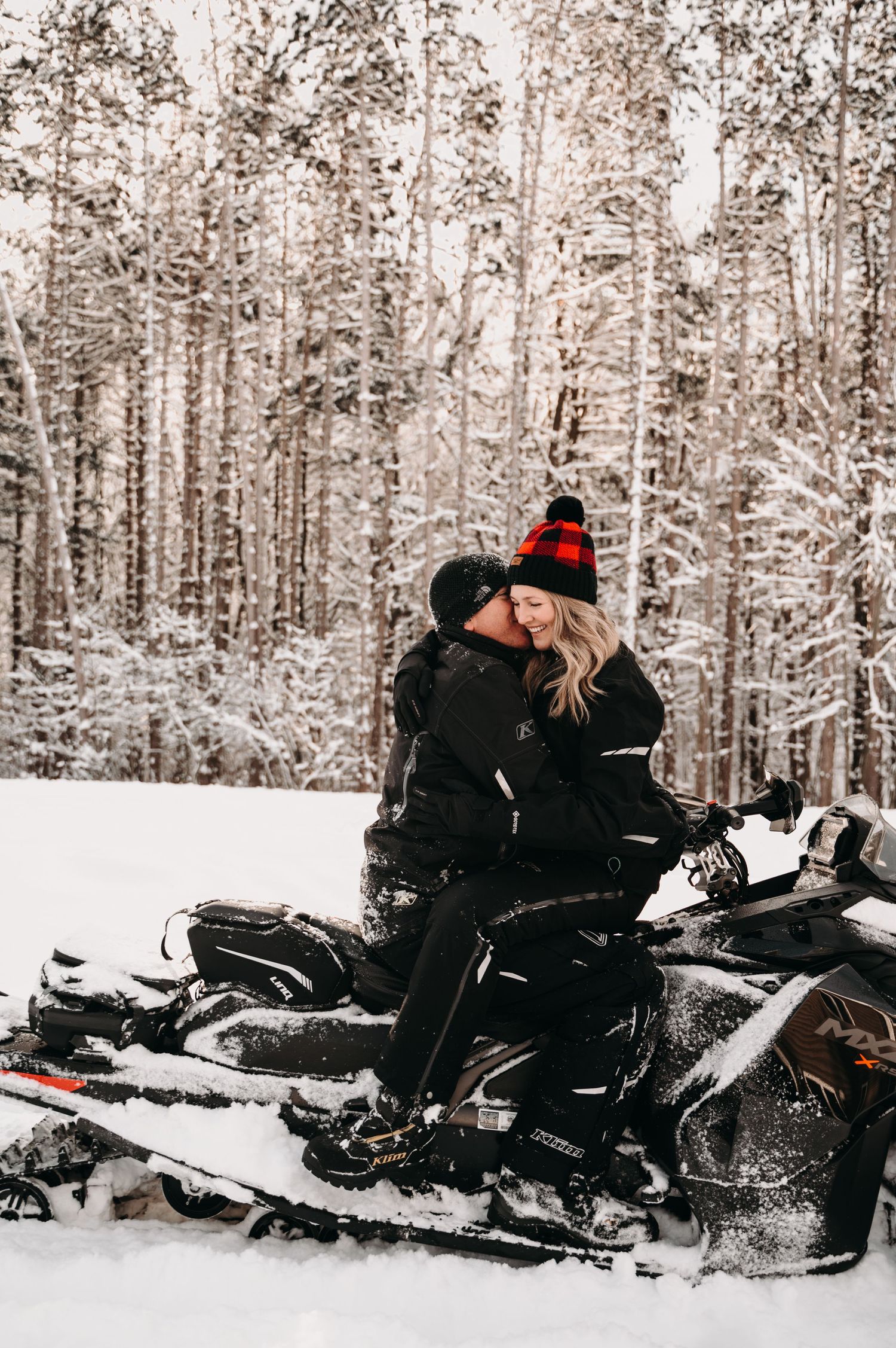 Couple sitting on snowmobile surrounded by snow-covered trees in winter forest.