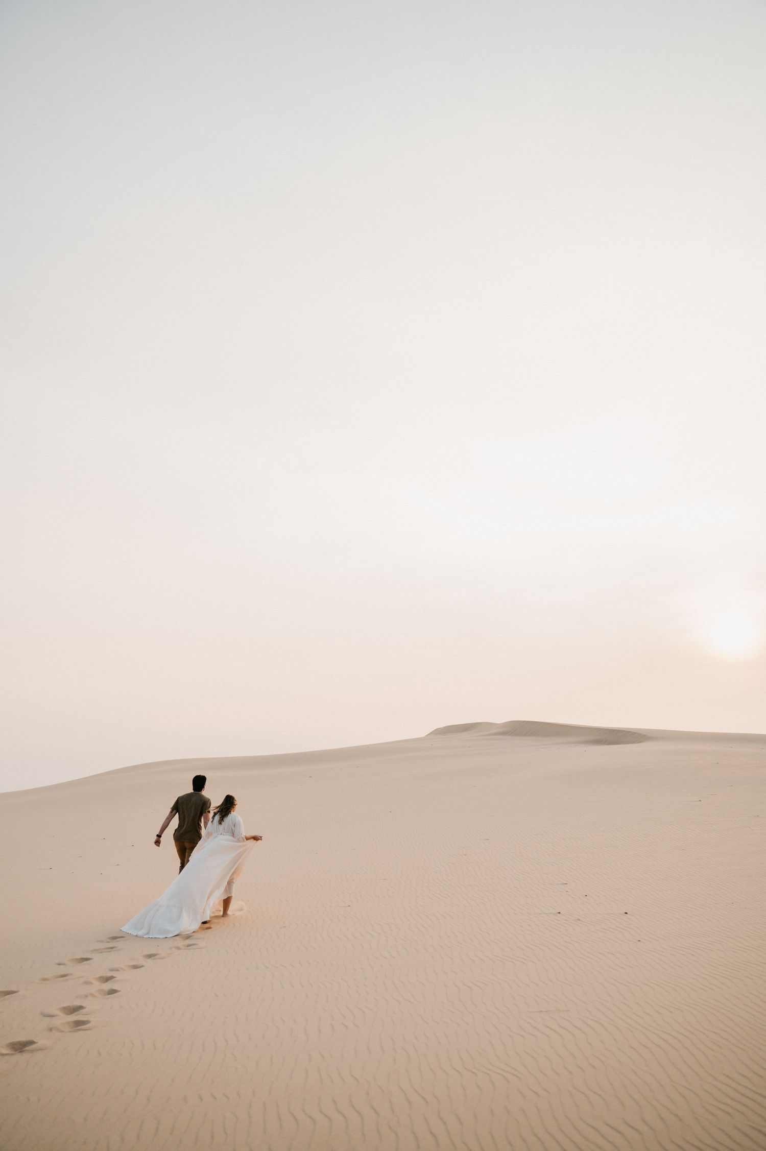 A couple walks hand in hand across desert sand dunes during golden hour.