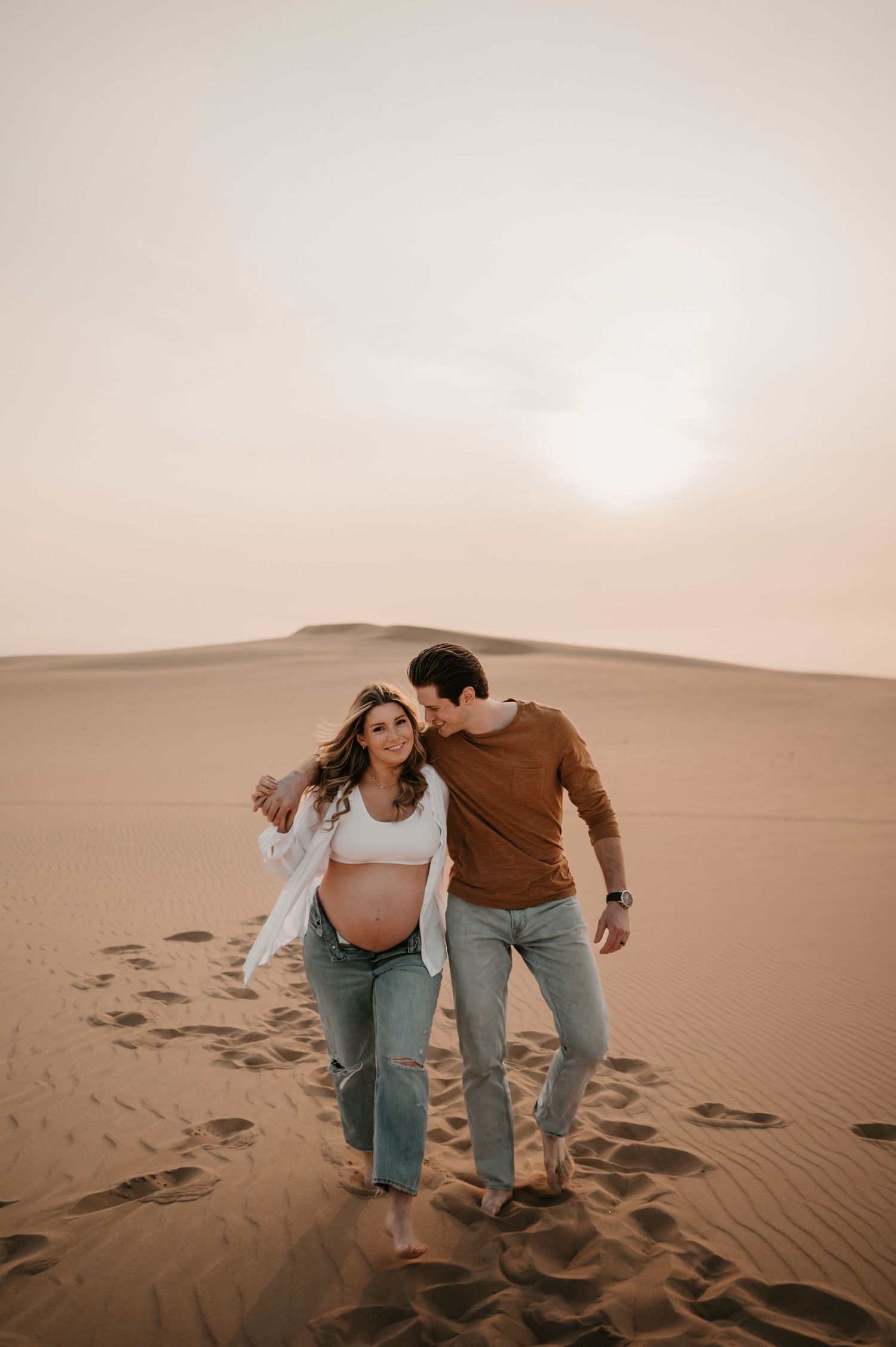 Romantic desert maternity photoshoot with couple walking through sand dunes.