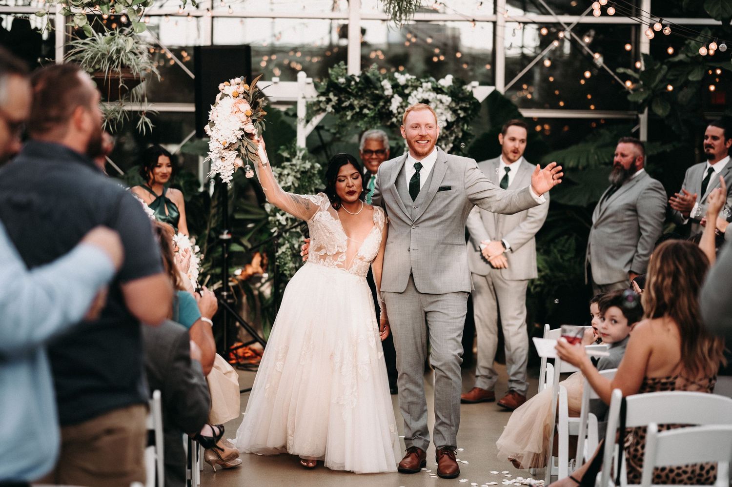 Newlyweds celebrate with raised arms while exiting down the aisle at greenhouse wedding.