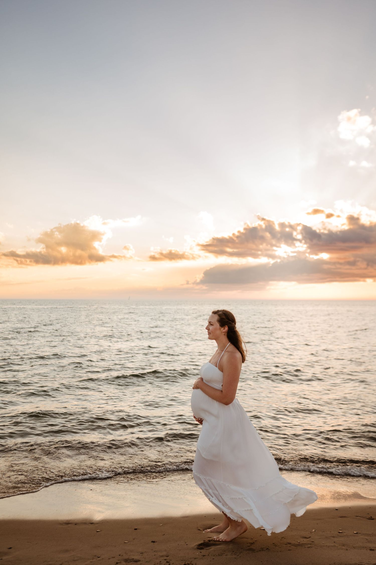 A pregnant woman in a flowing white dress walks along the beach at sunset while waves crash on the shore.