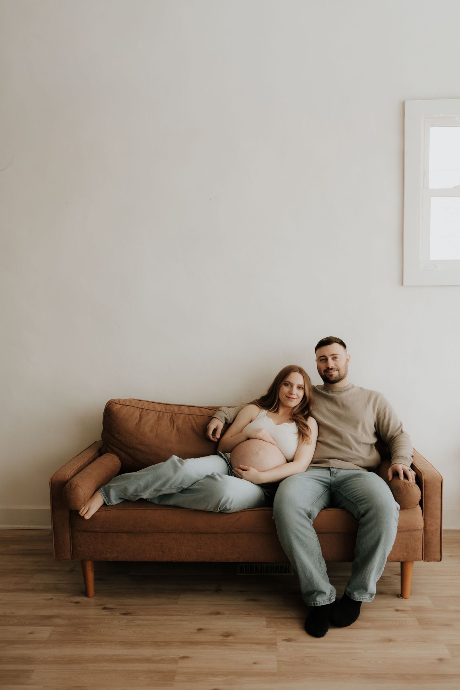 Relaxed maternity photo of couple lounging on brown leather couch in minimal setting.