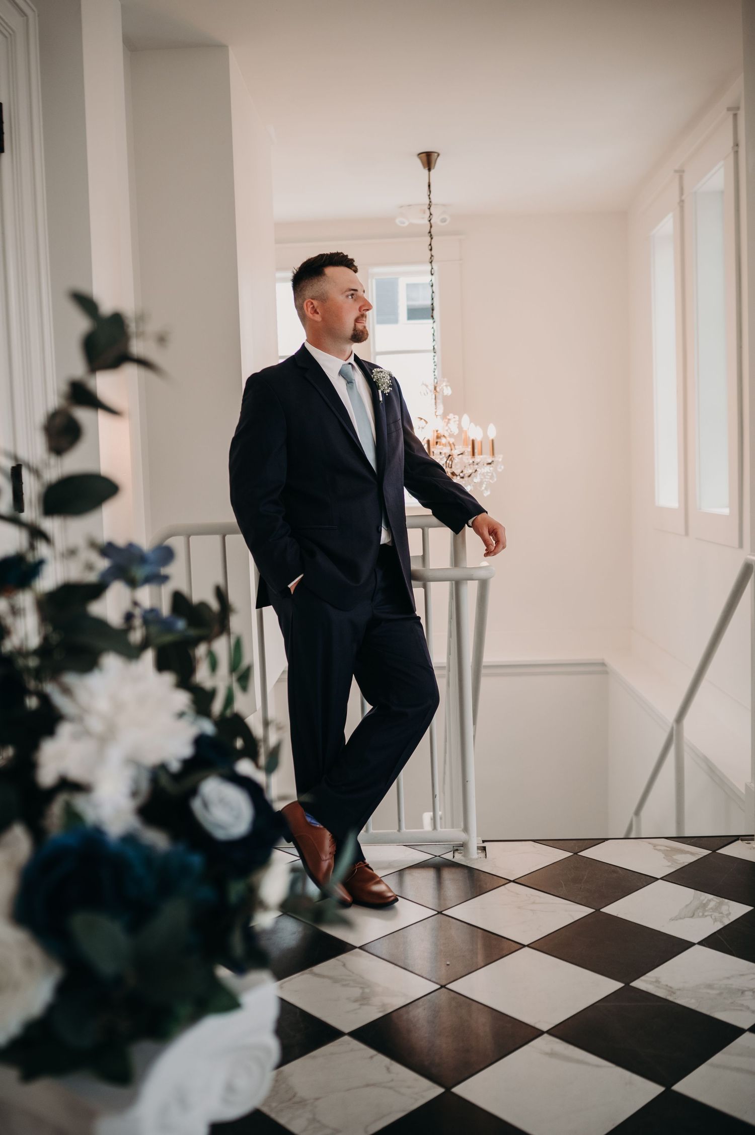 A person in a dark suit poses on checkered floor near floral arrangements and chandelier.