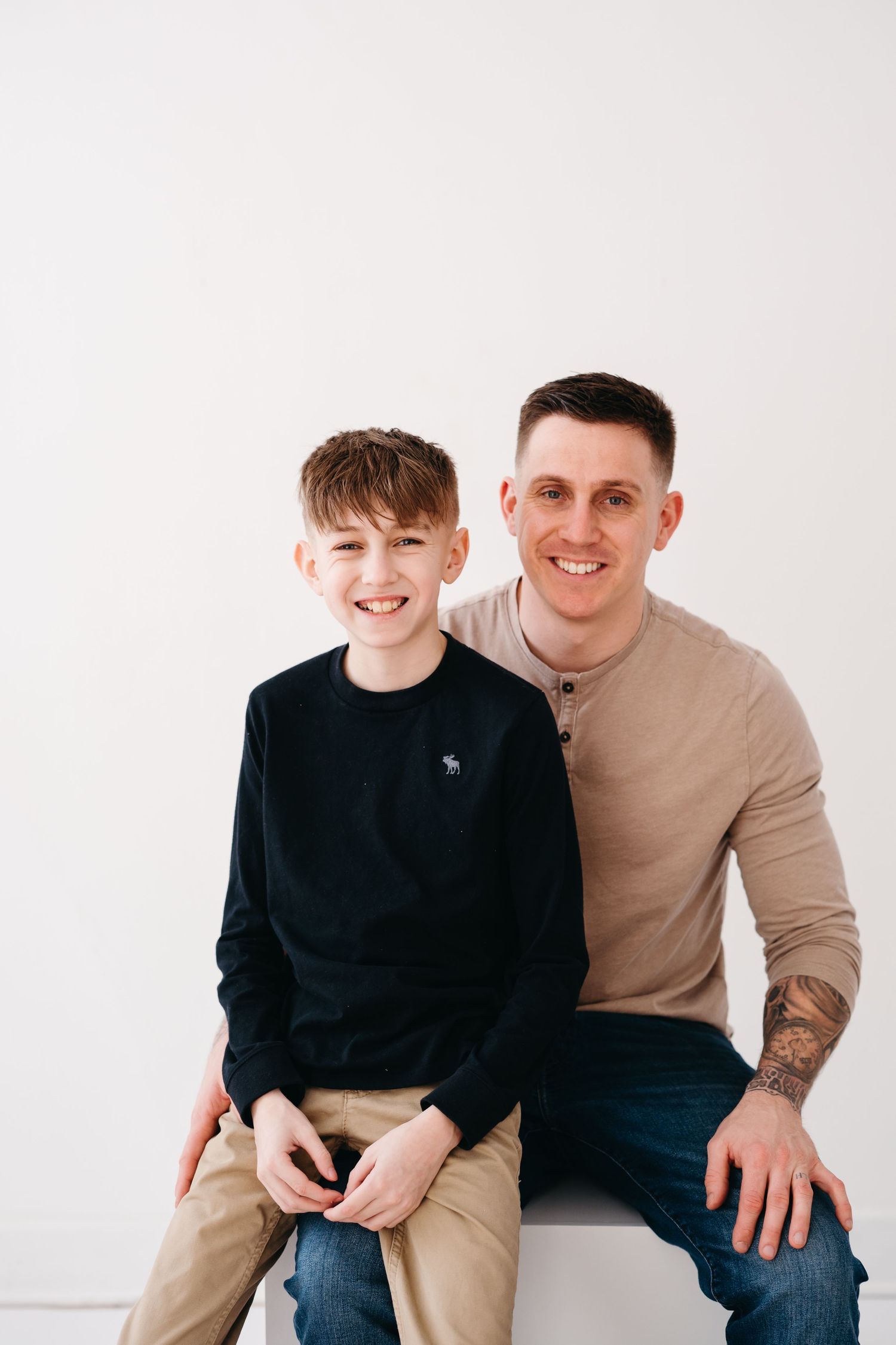 Two people sit together against a white background, wearing casual clothing and smiling.