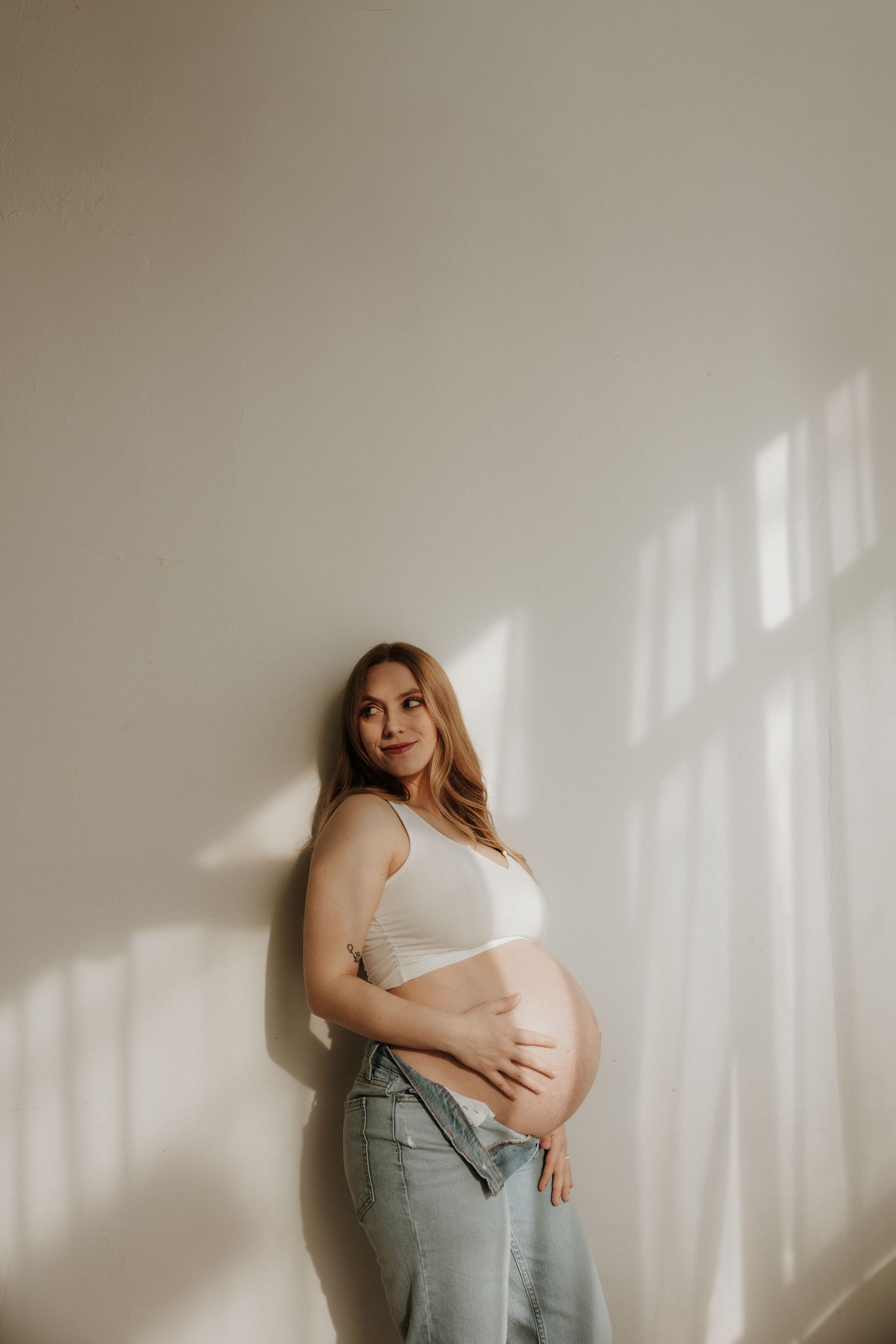 Maternity photoshoot in white top and jeans against neutral wall with soft window lighting.
