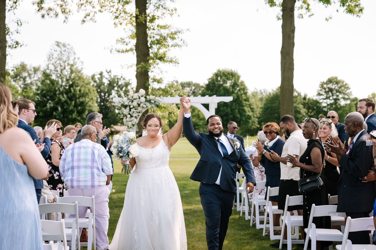Wedding ceremony procession on a grassy lawn with rows of white chairs and guests in attendance.