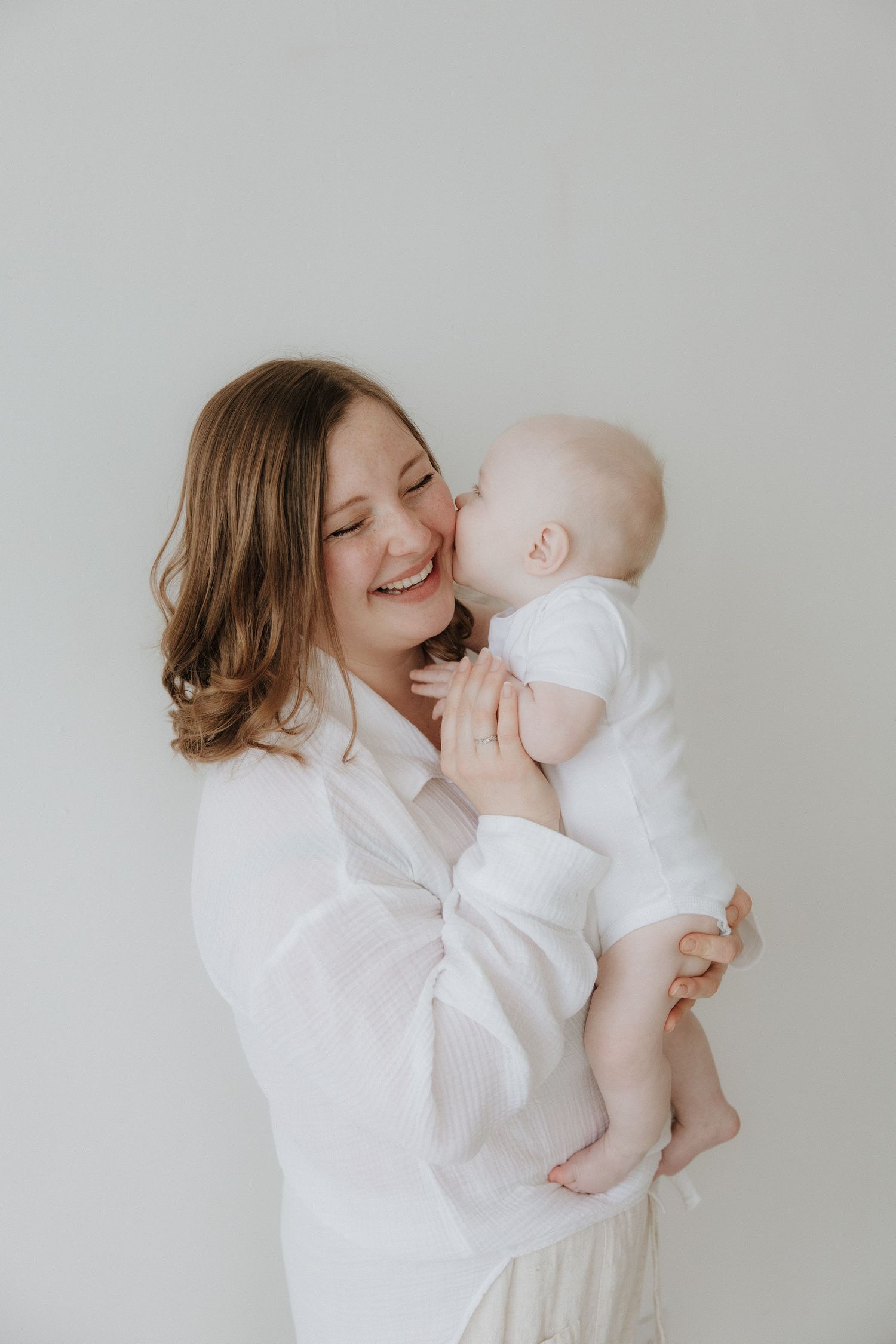 A joyful mother in a white blouse laughs as her baby gives her a sweet kiss on the cheek.