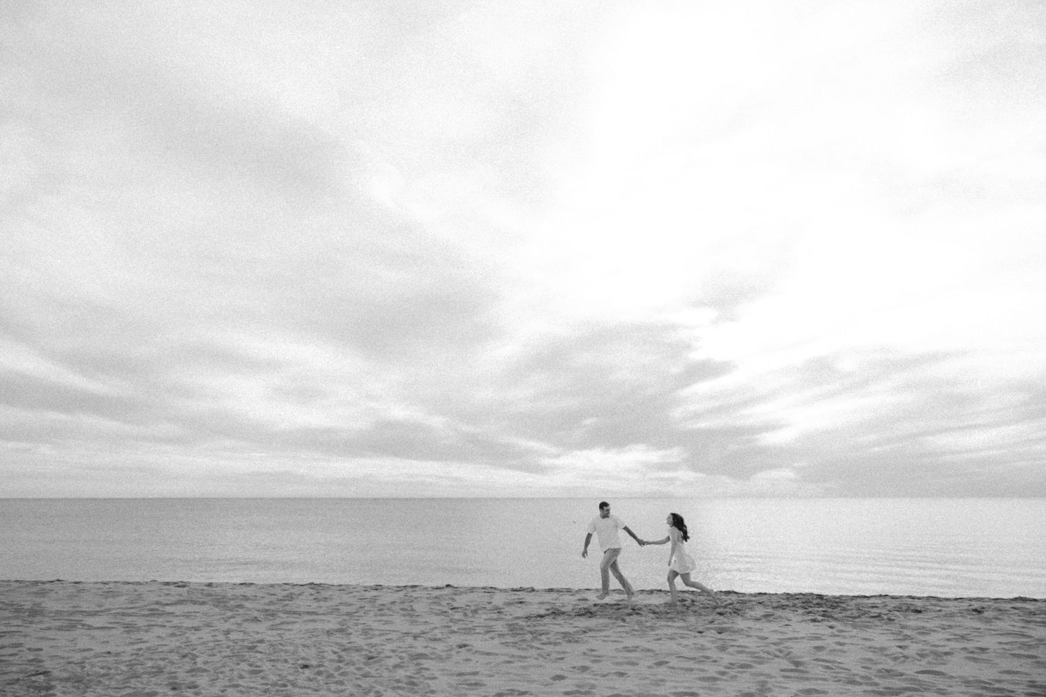 Black and white photo of a couple holding hands walking on a sandy beach under a cloudy sky.