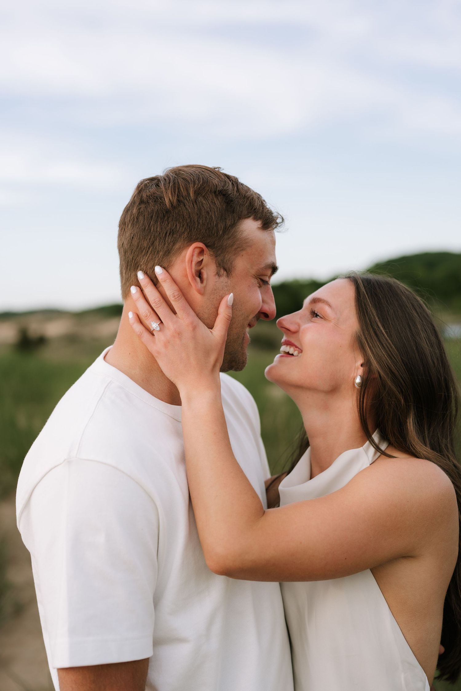Couple in white outfits sharing a romantic moment outdoors, woman smiling and touching mans face, engagement ring visible.