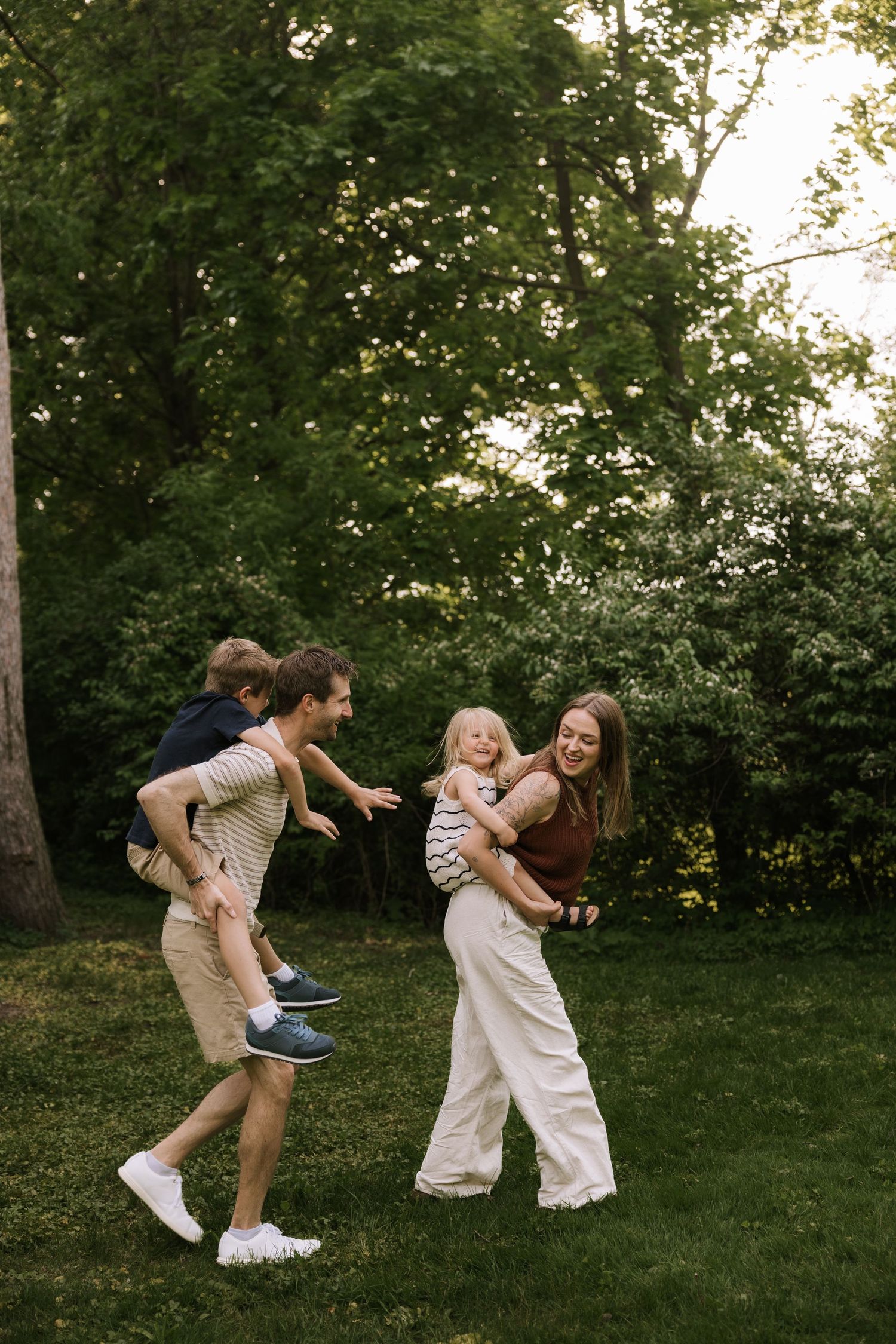 A happy family plays outdoors in a lush green park, parents each carrying a young child while laughing together.