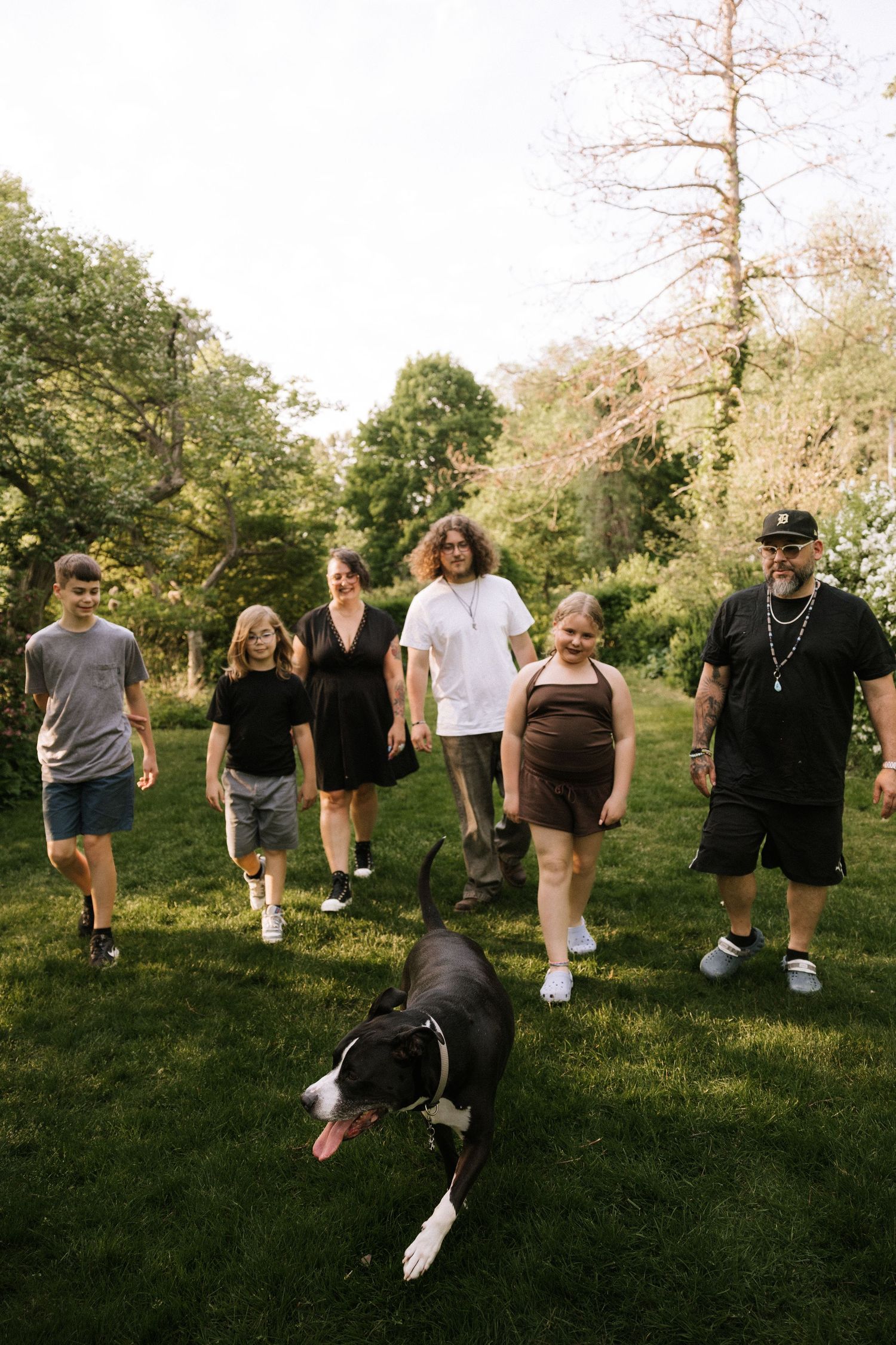 Family of six walking with black and white dog on lush green lawn surrounded by trees.