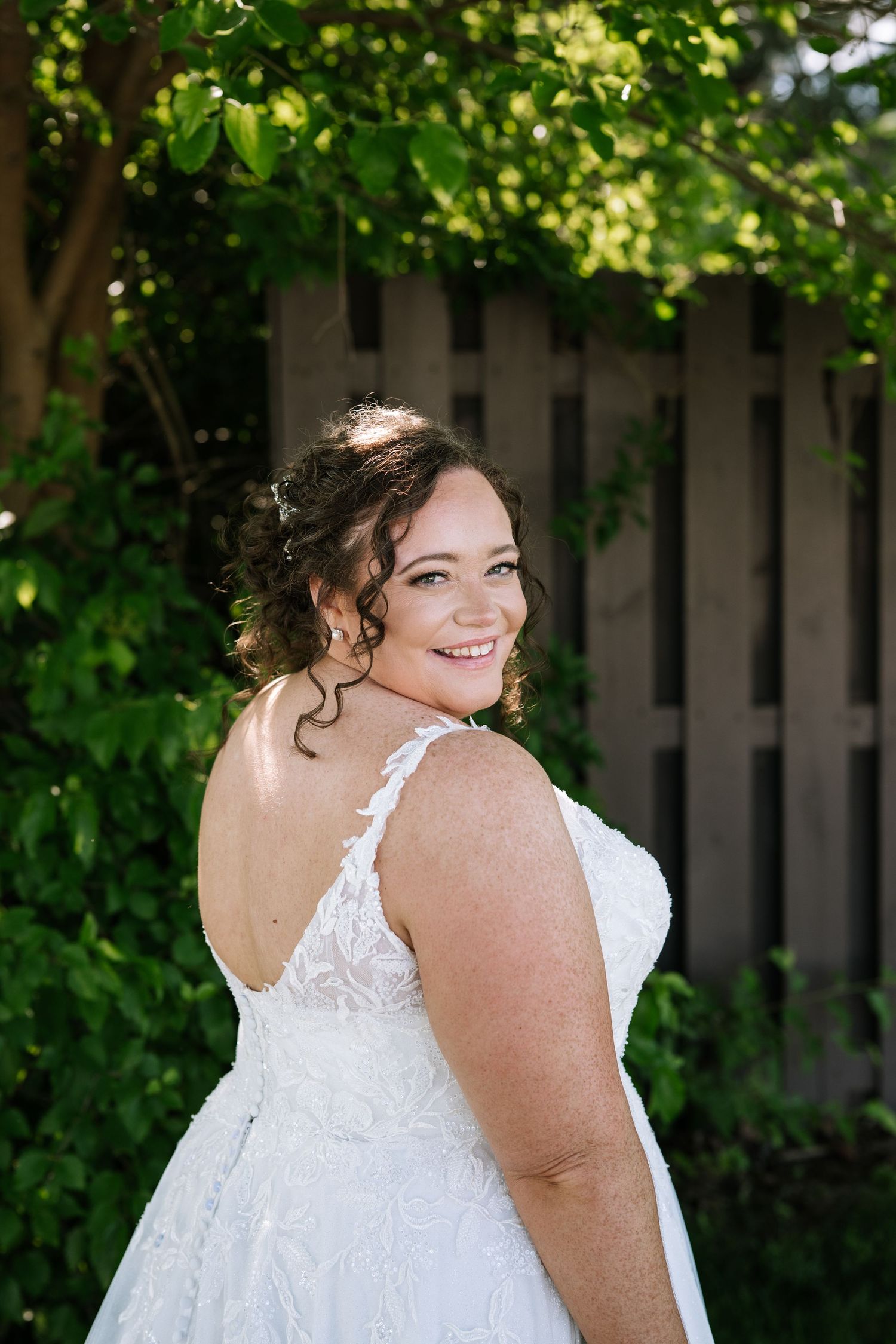 Bride in a white lace wedding dress poses outdoors against a wooden fence and green foliage backdrop.