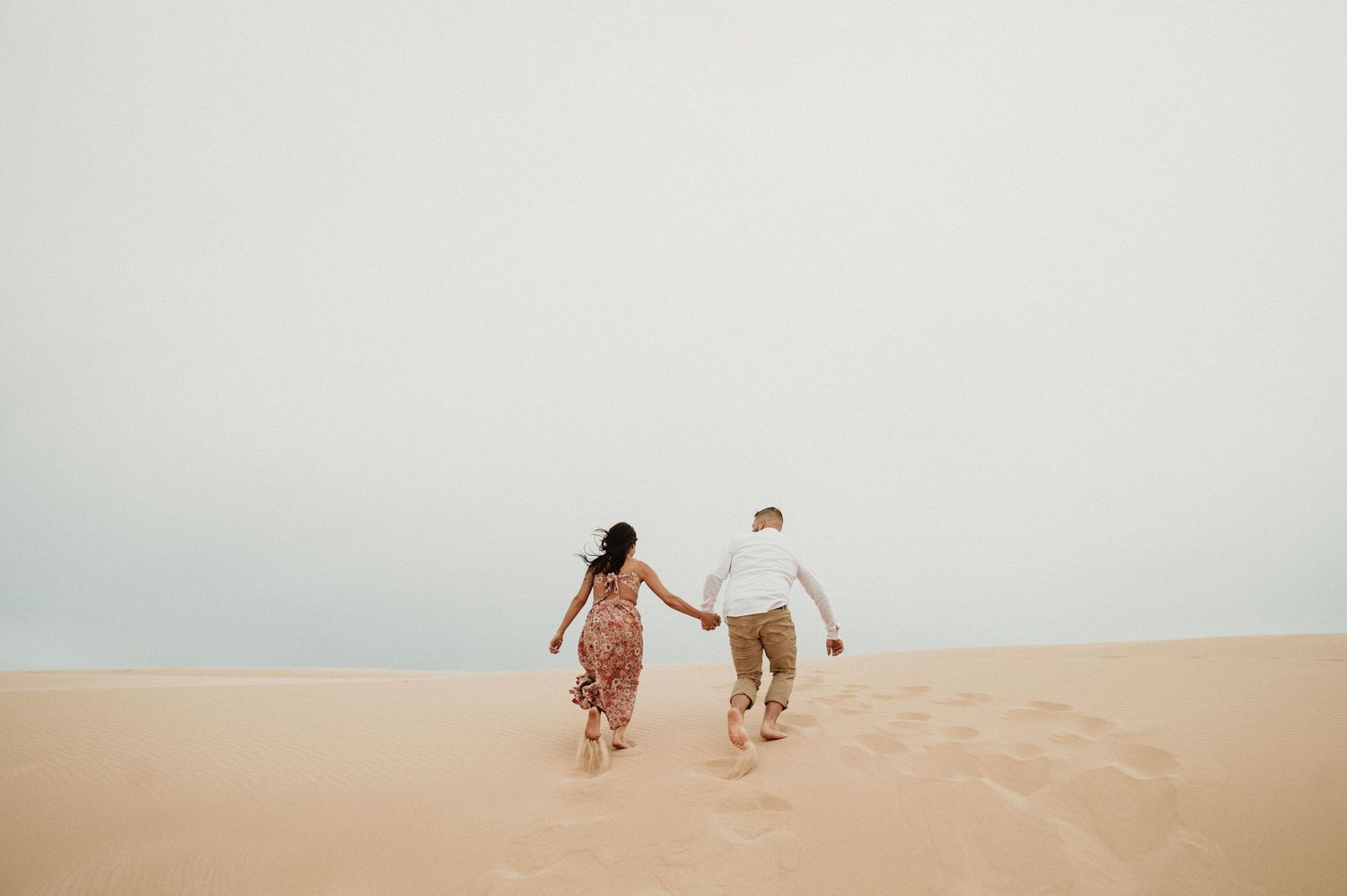 A couple runs playfully up a sand dune holding hands during sunset.