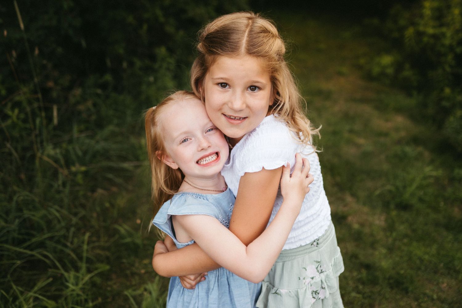 Two children share a loving embrace outdoors while wearing summer dresses.