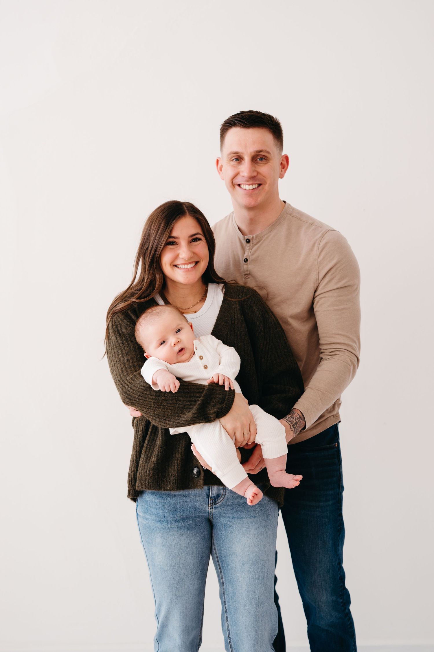 A couple stands together with their baby wearing a white outfit against a minimalist backdrop.